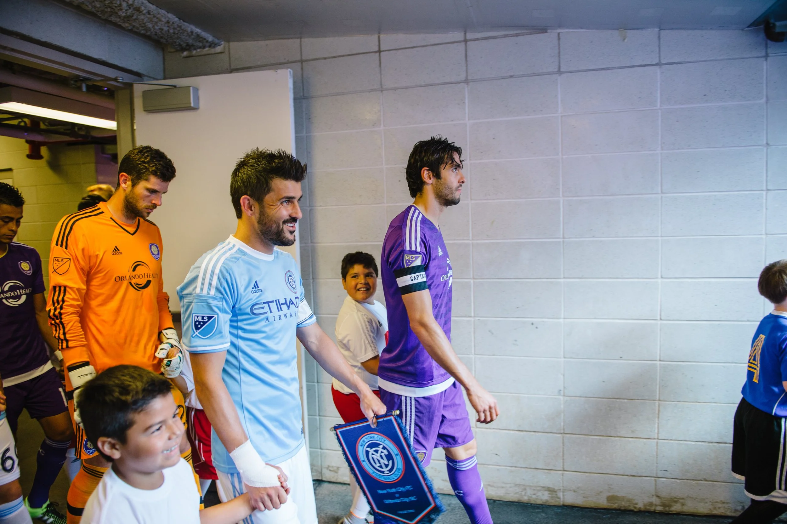 Soccer players walking through a corridor before a game, wearing different colored jerseys, with children and fans around.