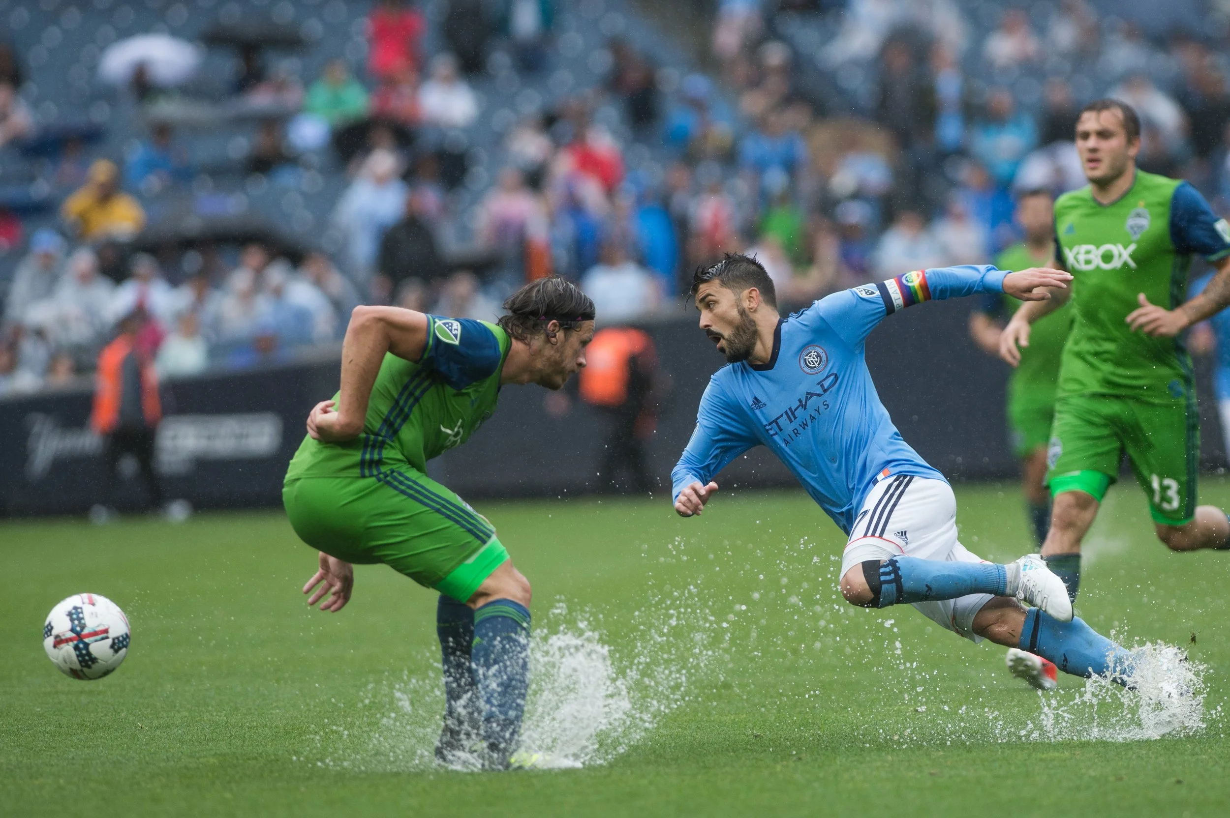 Two soccer players competing for the ball on a wet field, with spectators in the background. One player wears a blue jersey and shorts with the Etihad Airways logo, and the other in a green jersey and shorts. It is raining.
