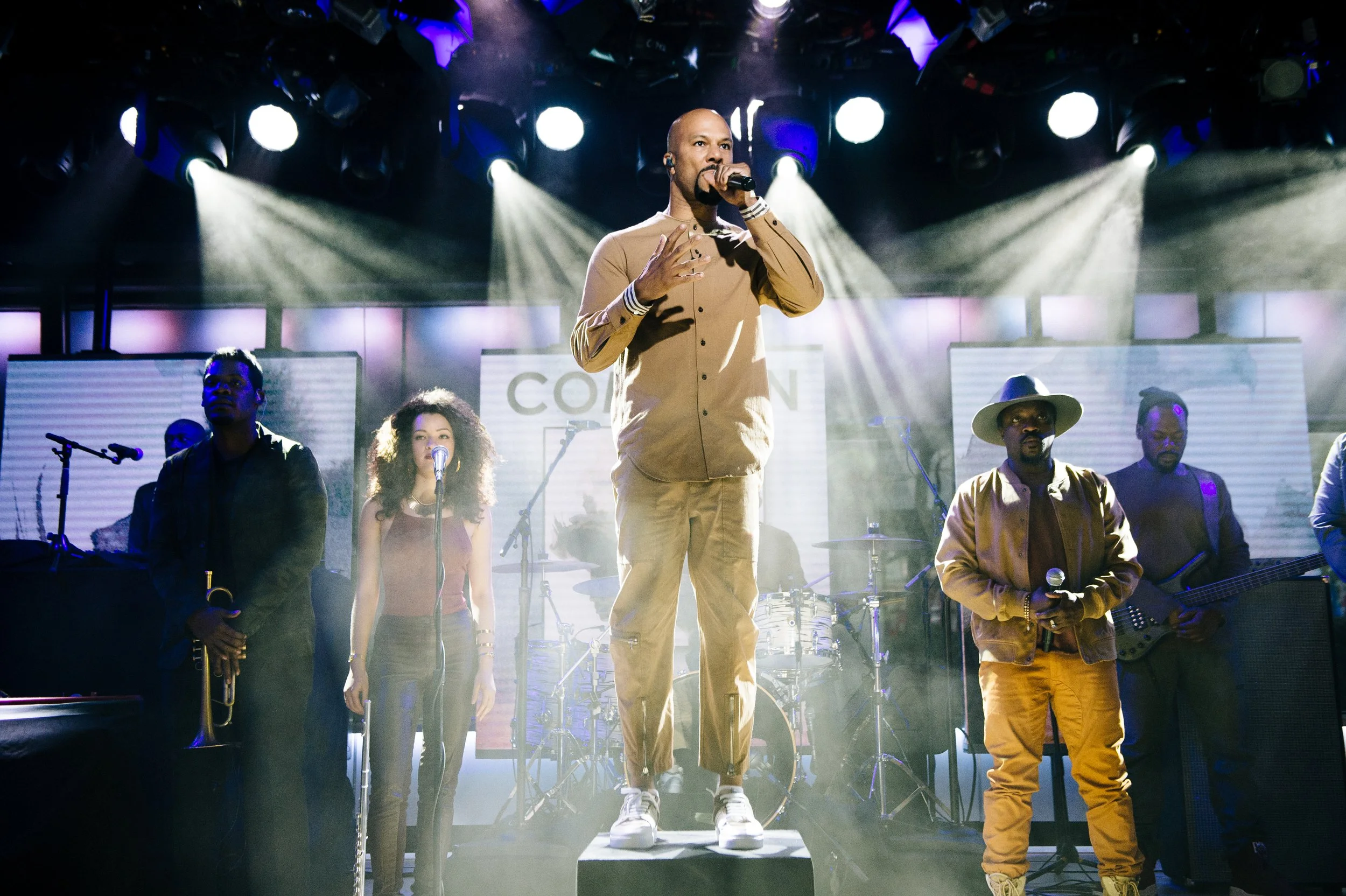 Musicians performing on stage with bright lights shining down, including a male singer in the center wearing a beige outfit and sneakers, surrounded by band members playing instruments and singing.