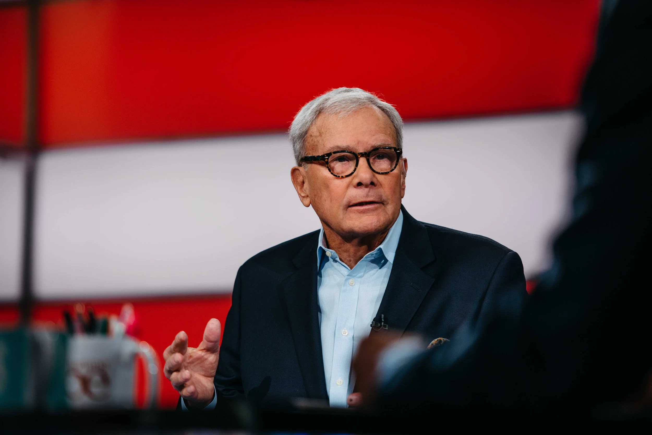 A man with gray hair and glasses wearing a blue shirt and dark blazer, speaking during an interview or discussion, with a blurred person in the foreground and a red, white, and gray background.