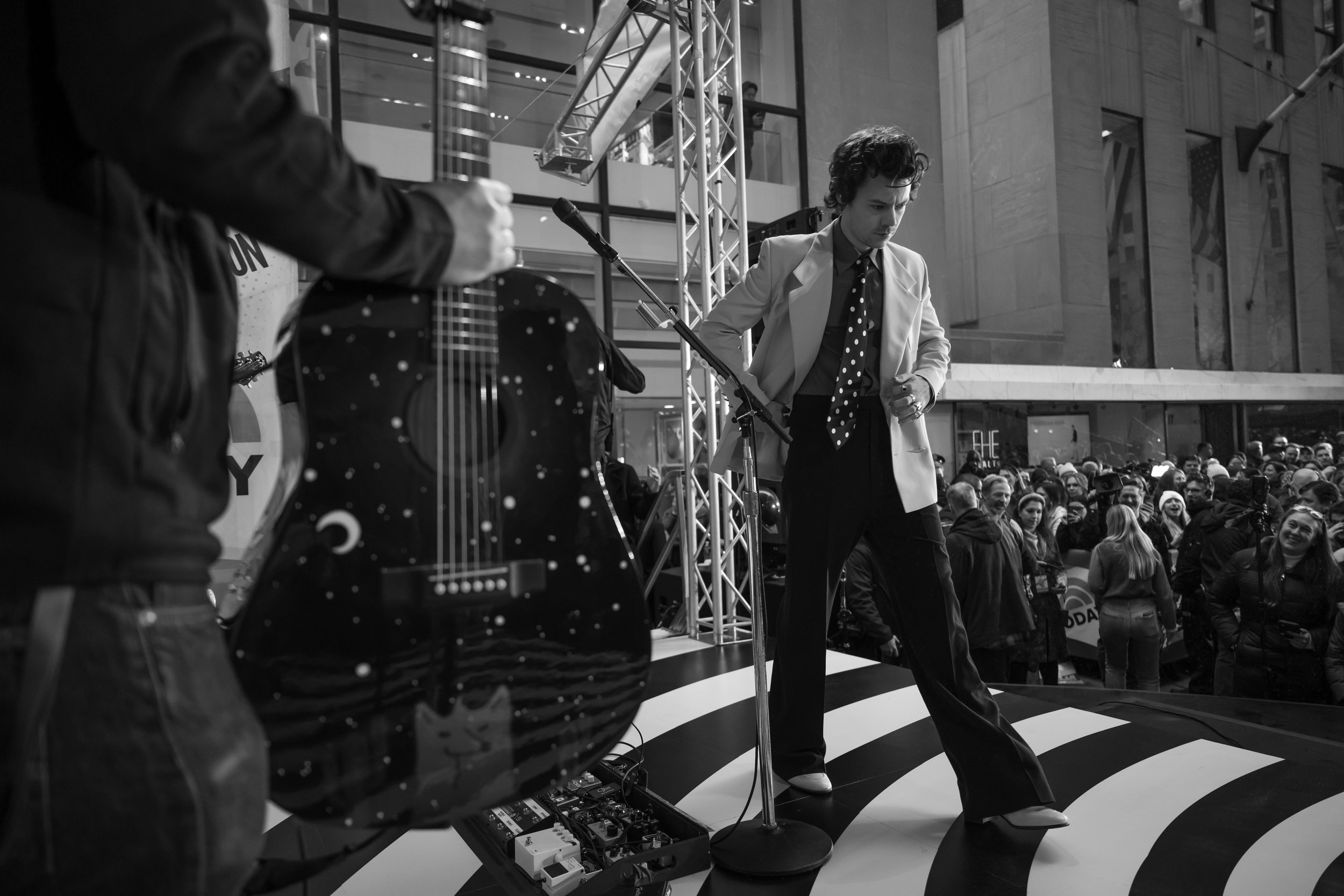 A black and white photo of a performer on a stage with a crowd watching. The performer has curly hair, is dressed in a light blazer, dark pants, and a polka dot tie, and is looking down. A guitar with a cosmic design is visible in the foreground.