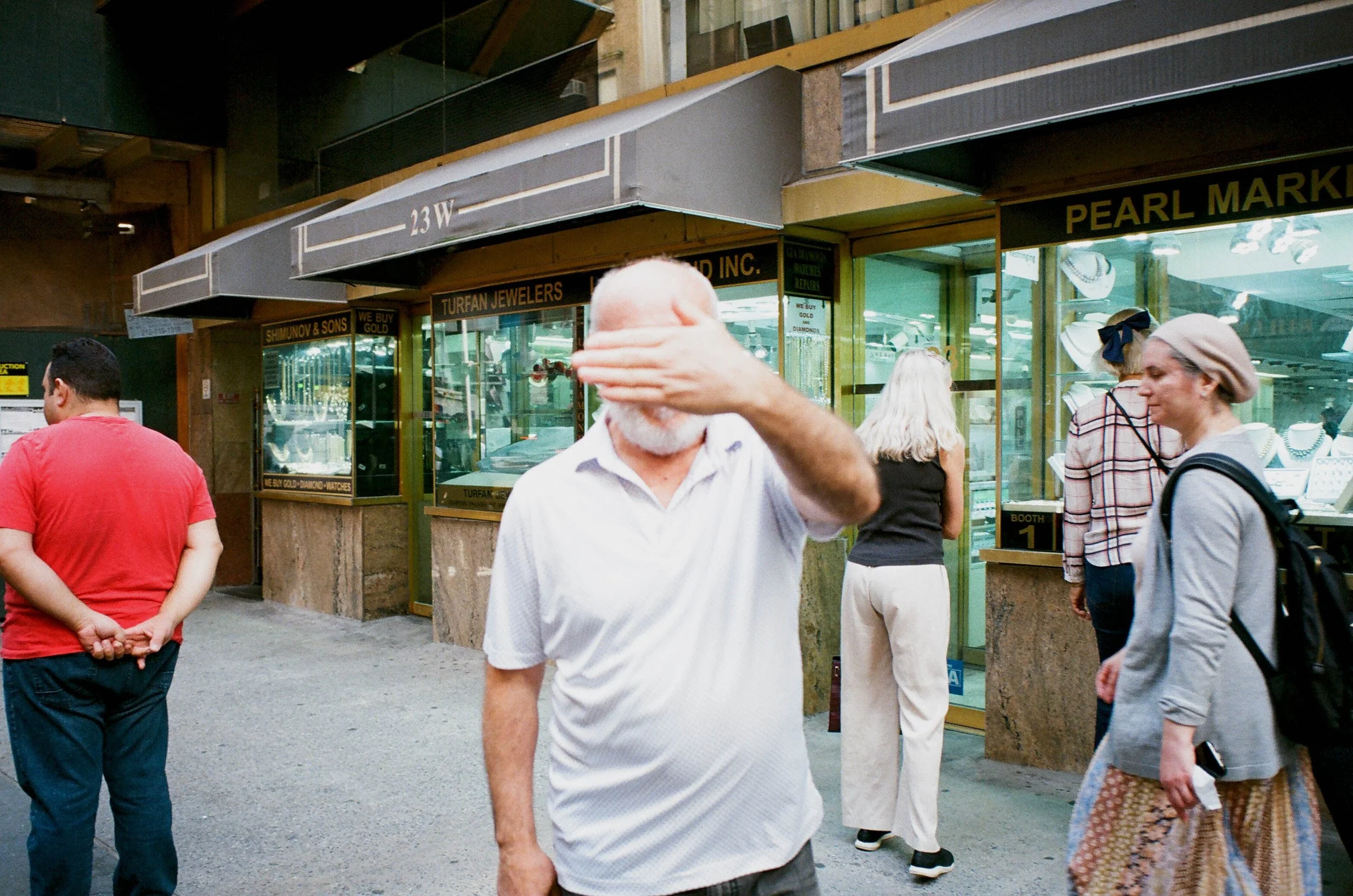 A man with white hair and a beard in a white shirt is covering his face with his hand, standing outside a jewelry store with other pedestrians nearby.