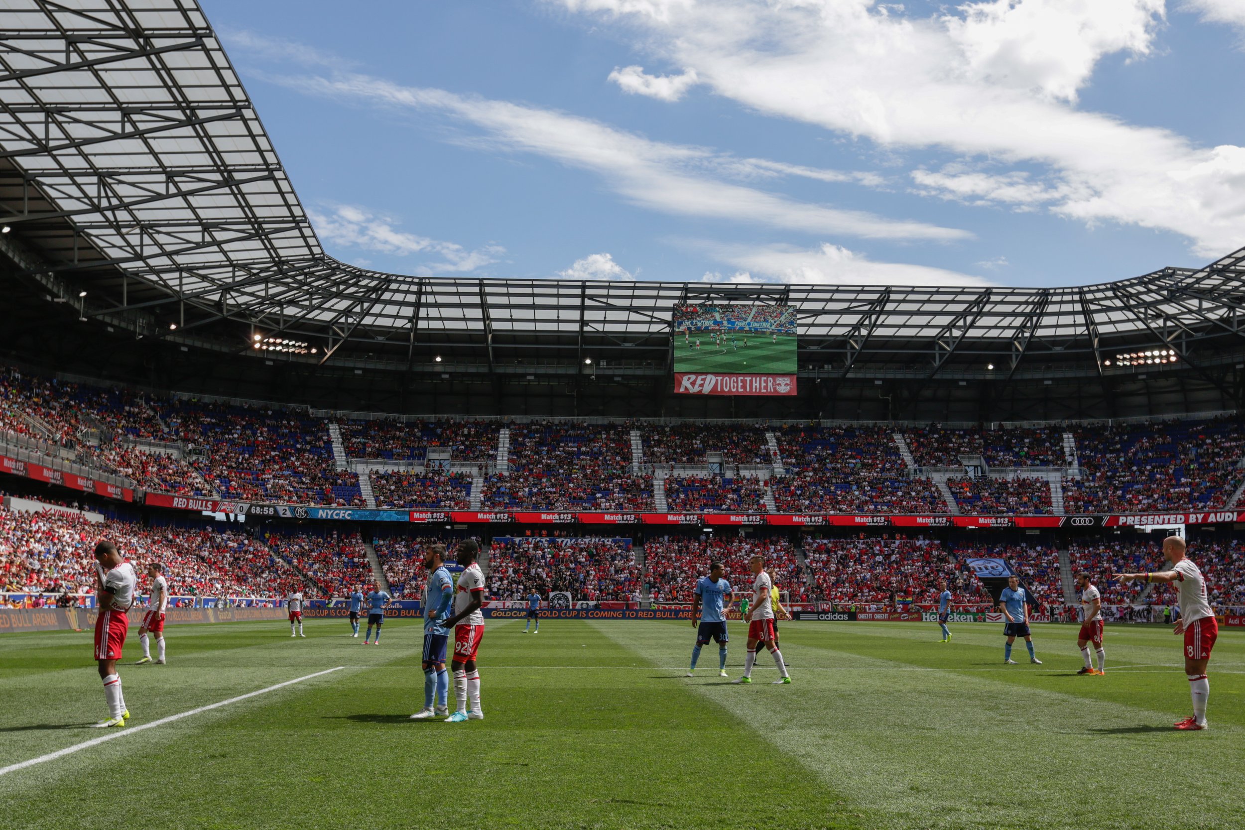 Soccer players on the field during a game at a large stadium with a partially open roof, filled with spectators, under a partly cloudy sky.
