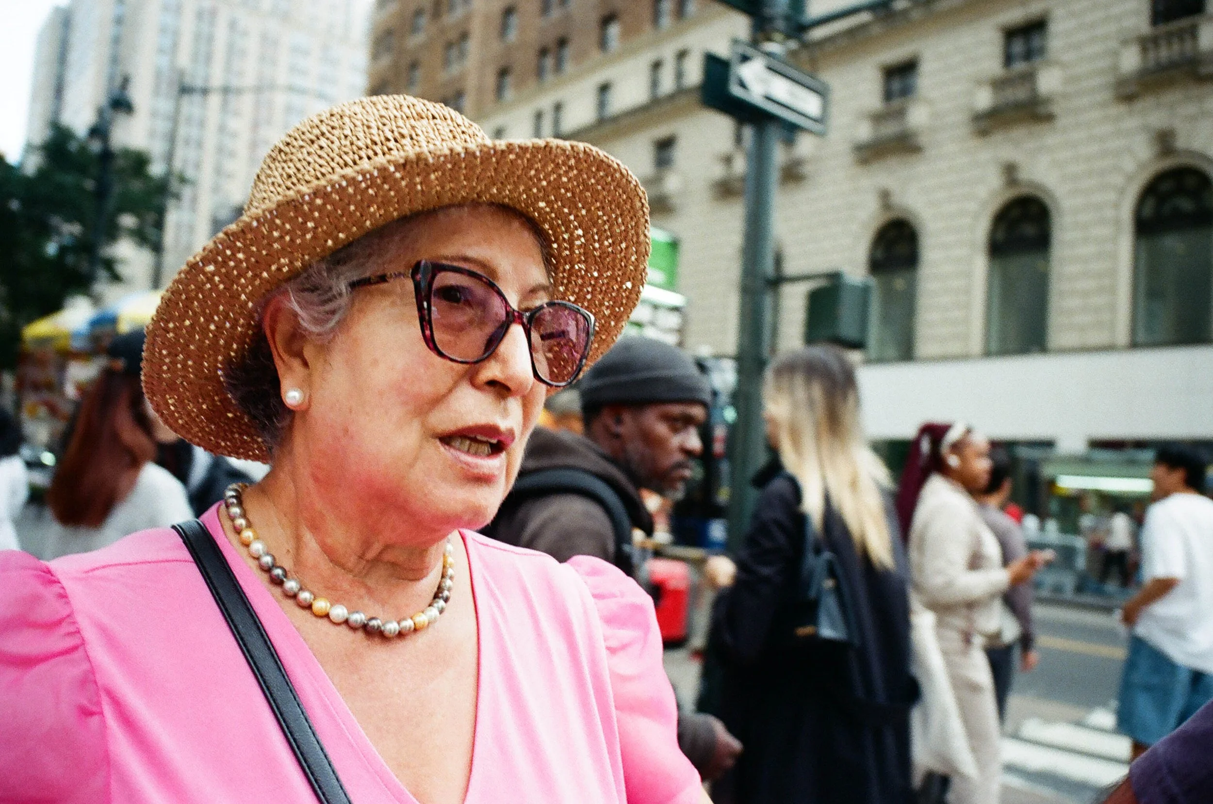 An elderly woman with short gray hair, wearing a pink blouse, pearl necklace, pearl earrings, large sunglasses, and a straw hat, walking on a city sidewalk with a crowd of people in the background.