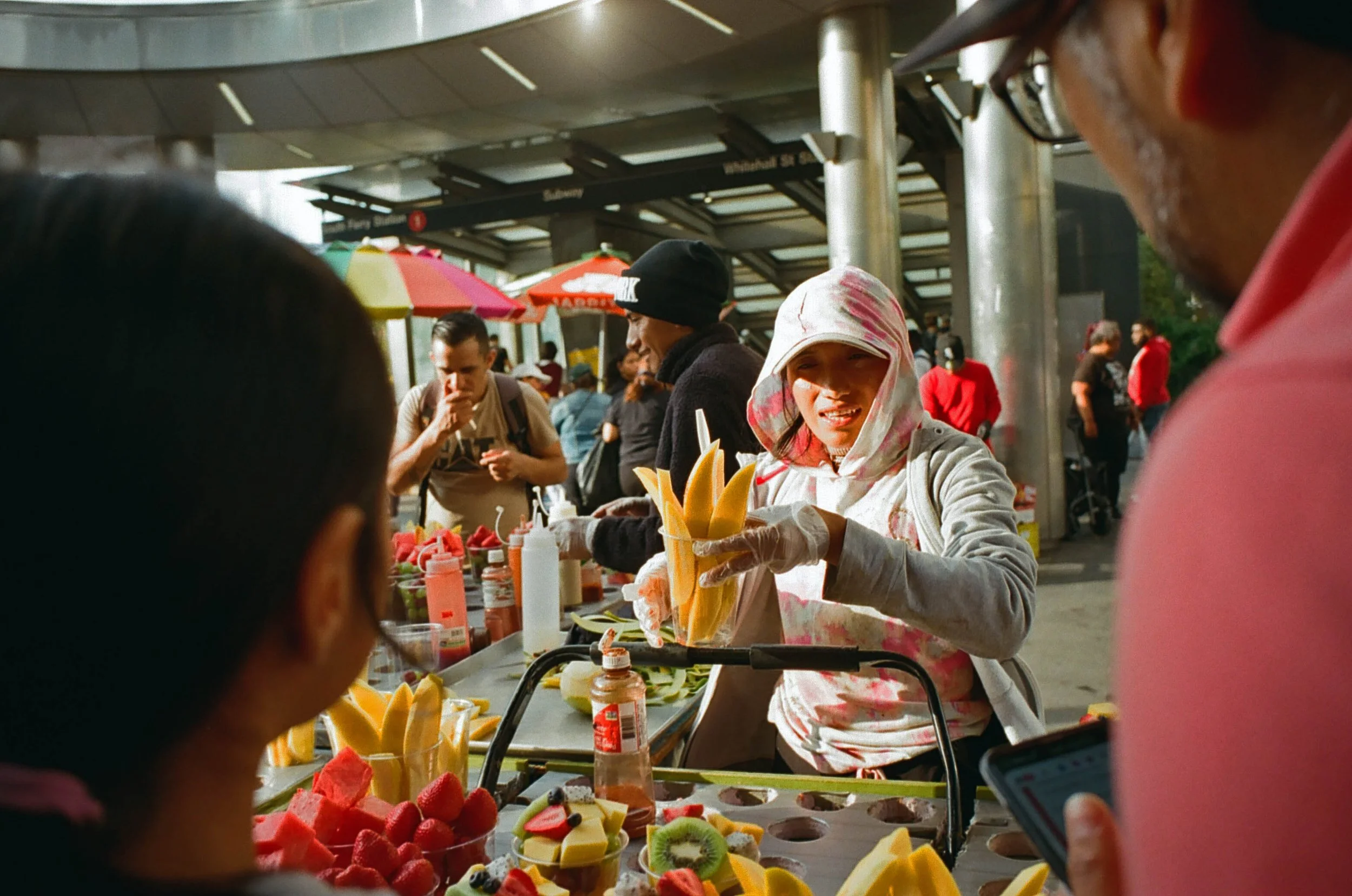 A street vendor selling sliced mangoes at a busy outdoor market under colorful umbrellas with people shopping and walking around in the background.