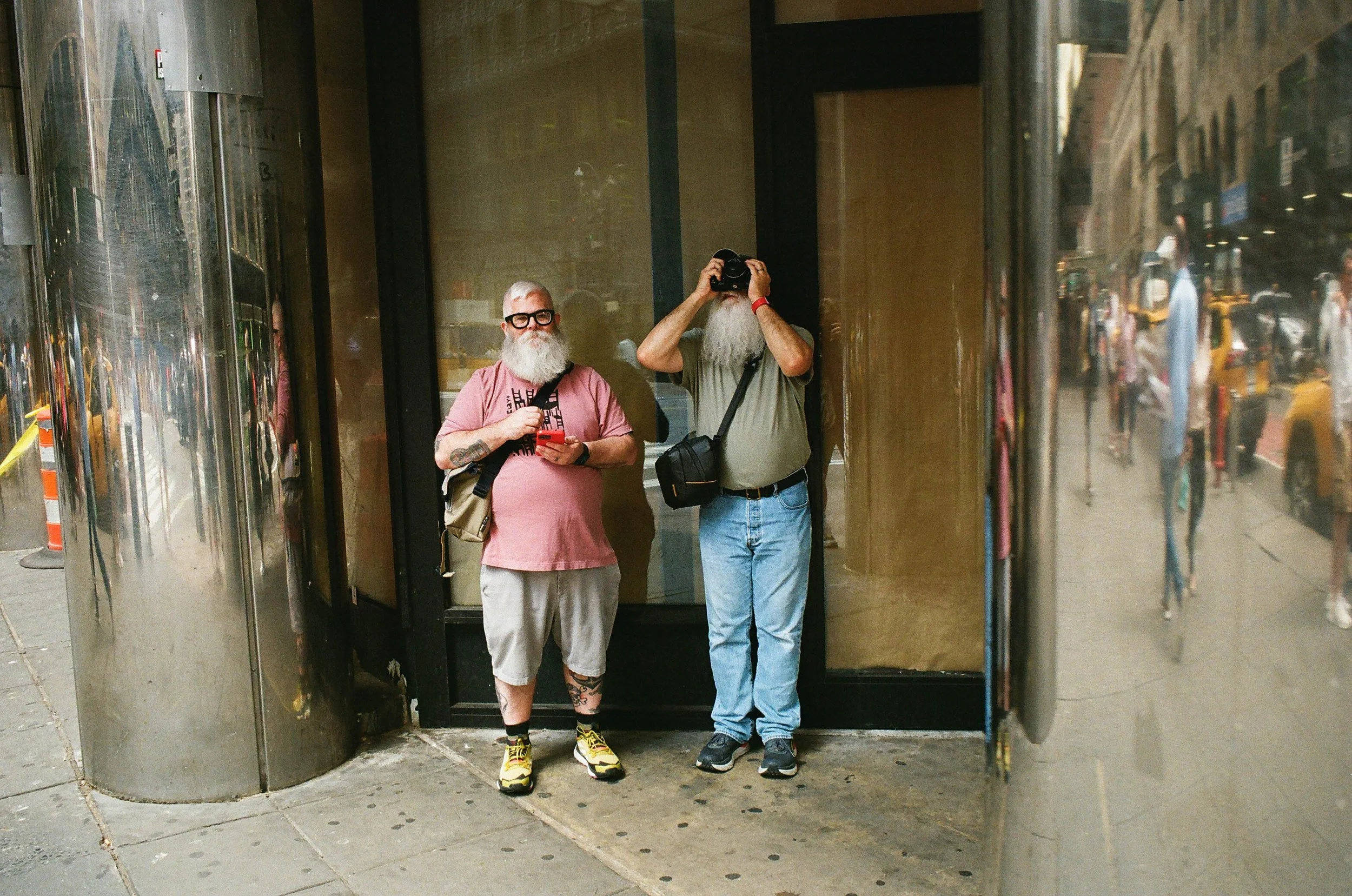 Two men with beards and casual clothing stand on a city sidewalk; one with glasses, holding a phone, the other with a camera.
