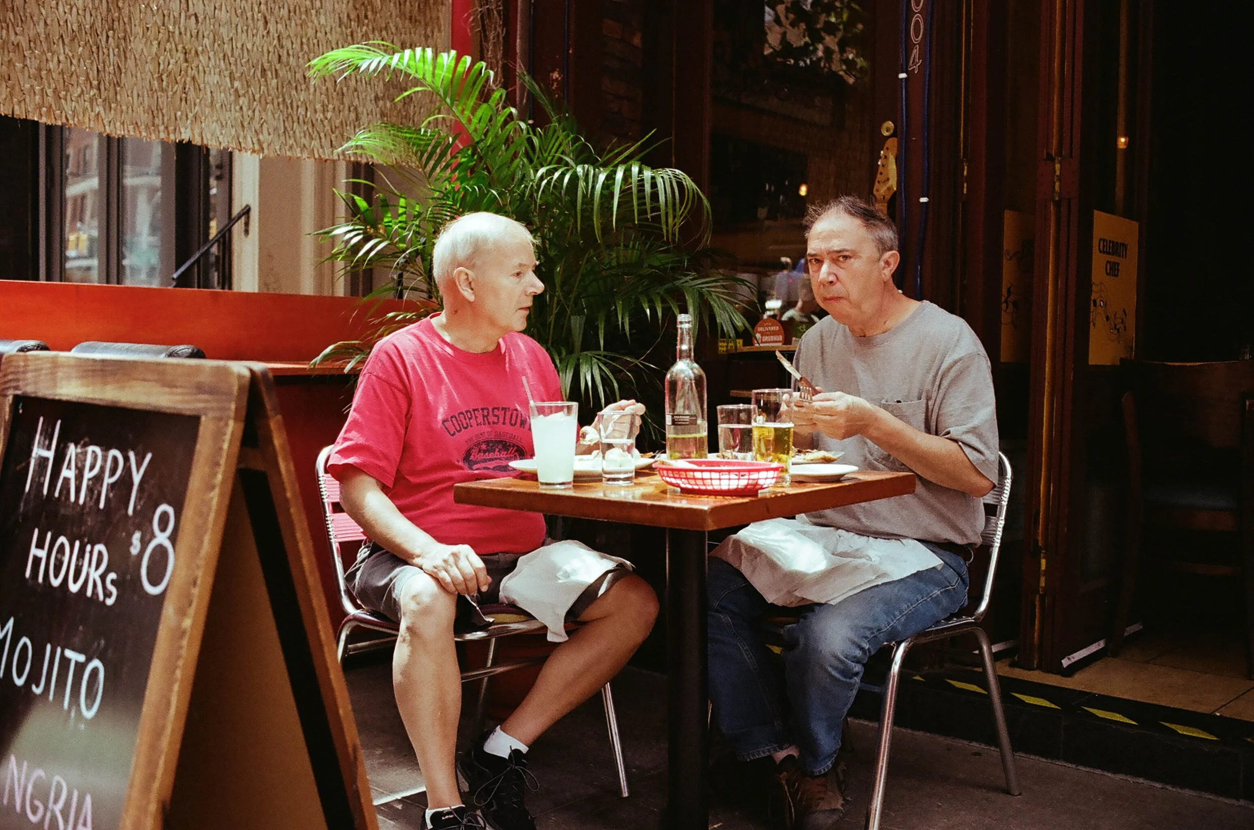 Two people sitting at a small table in a restaurant, having a conversation, with drinks and a bottle on the table, with an indoor plant and a window in the background.