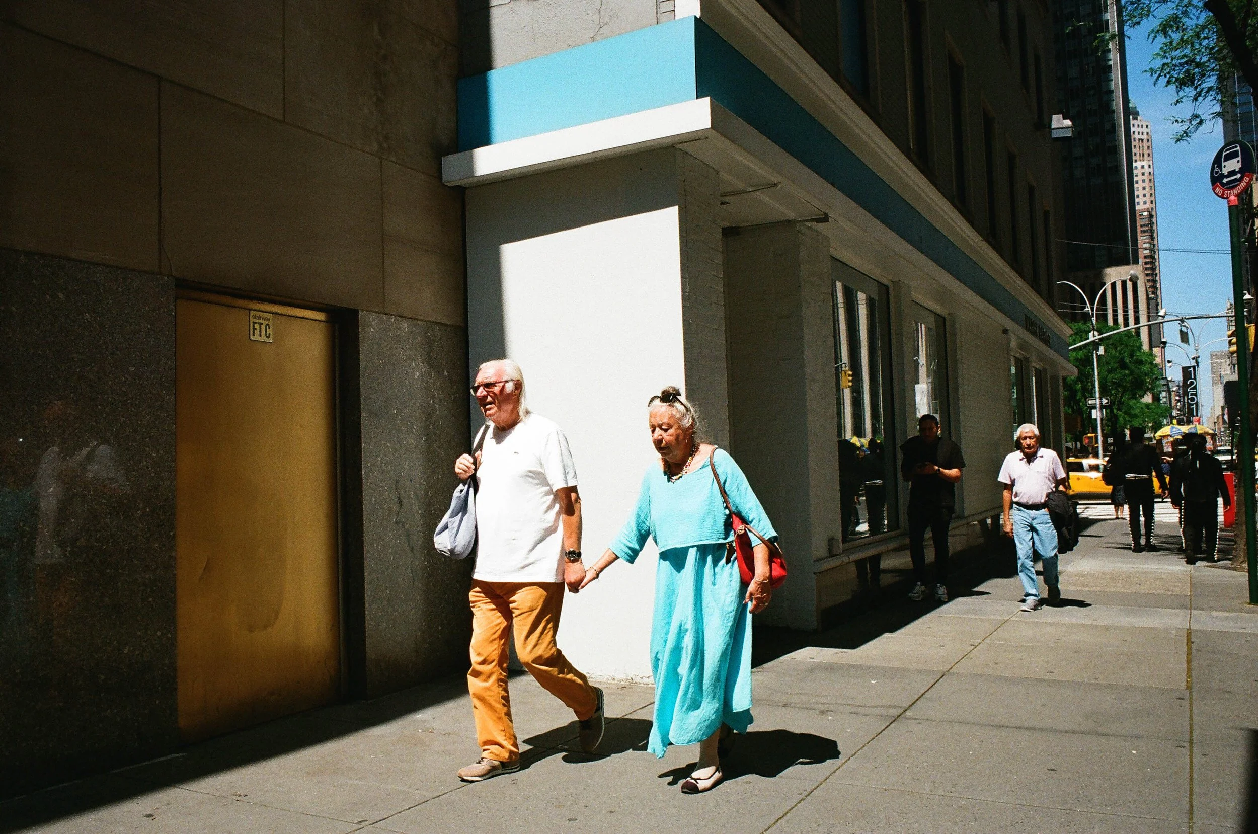 An elderly man and woman holding hands walking on a city sidewalk. The man is wearing a white shirt and light brown pants, carrying a gray bag. The woman is dressed in a light blue dress with white high-heeled shoes, carrying a red bag. Several other