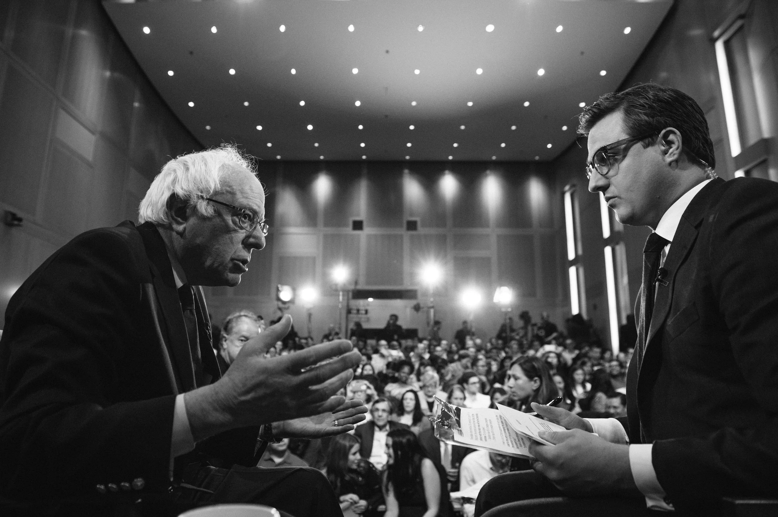 Two men in suits and glasses engaged in conversation at a large conference or seminar, with an audience seated in the background.