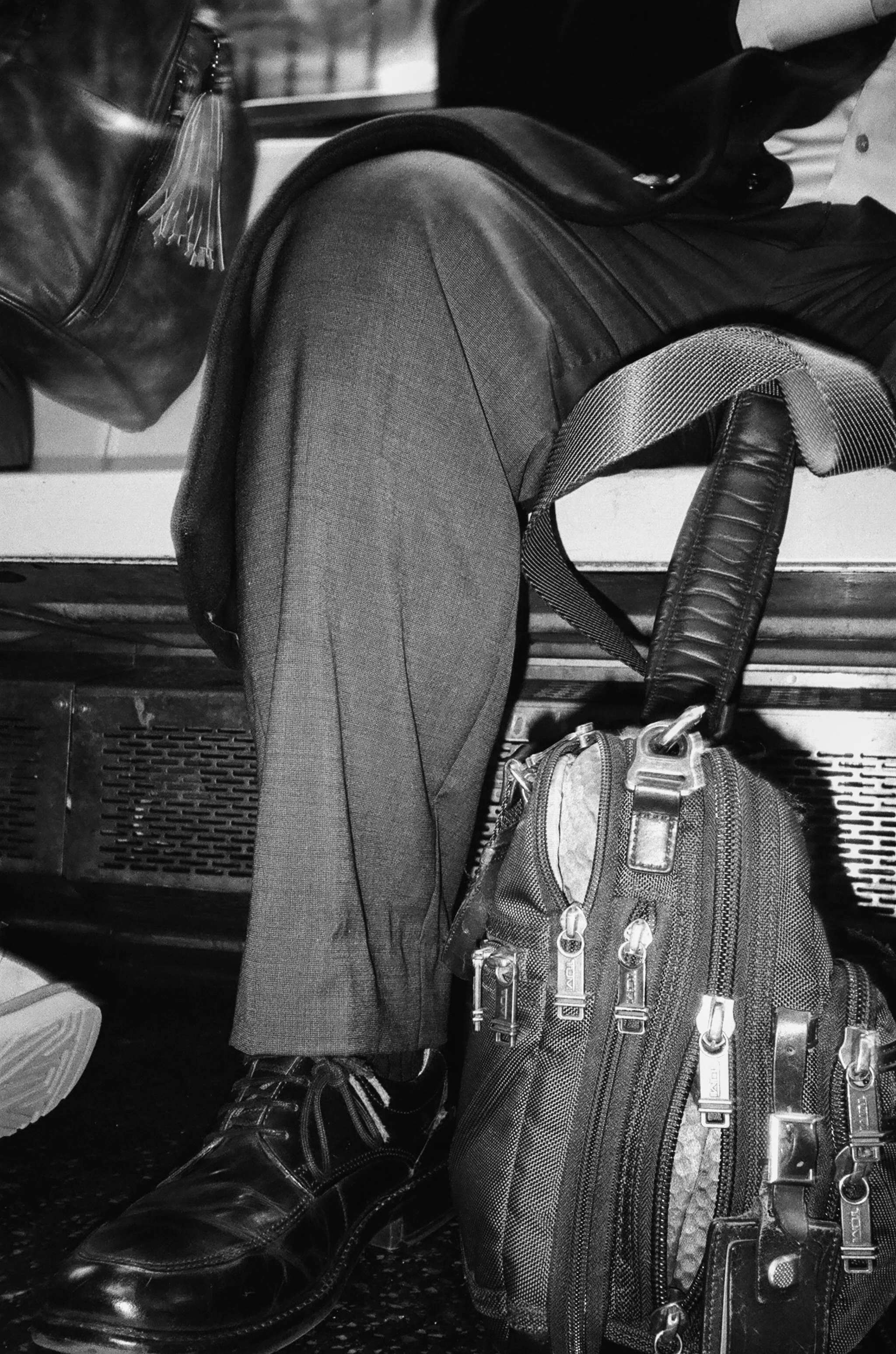 A person sitting with crossed legs, wearing dress shoes, dark trousers, and a dark jacket. A backpack is leaning against their leg, and a bag is placed nearby, with a leather bag visible in the upper left corner.