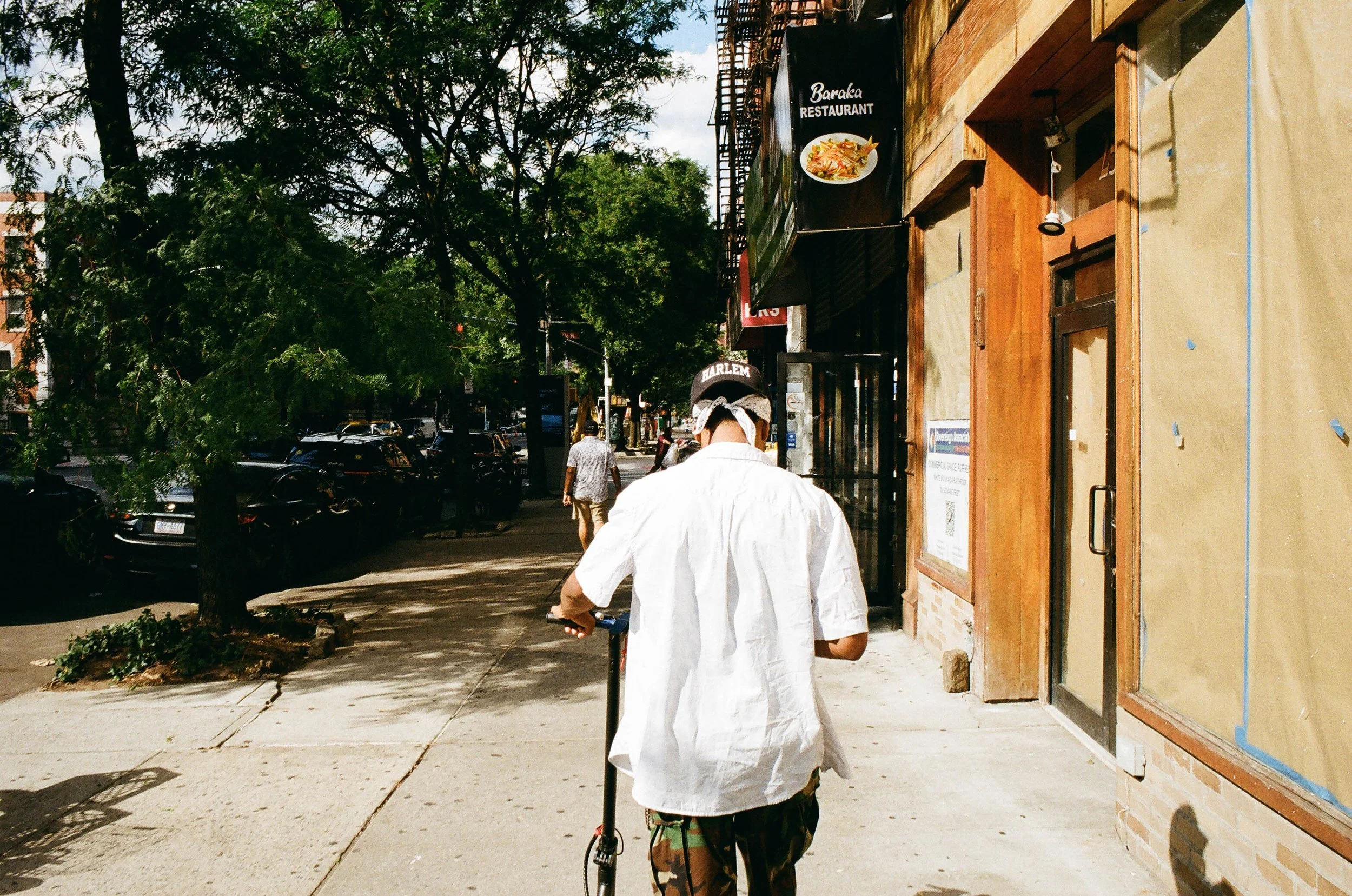 A man in a white shirt and camouflage pants riding a scooter on a sidewalk in an urban area. There are trees, parked cars, and pedestrians in the background, along with storefronts and signs, including one for a restaurant.