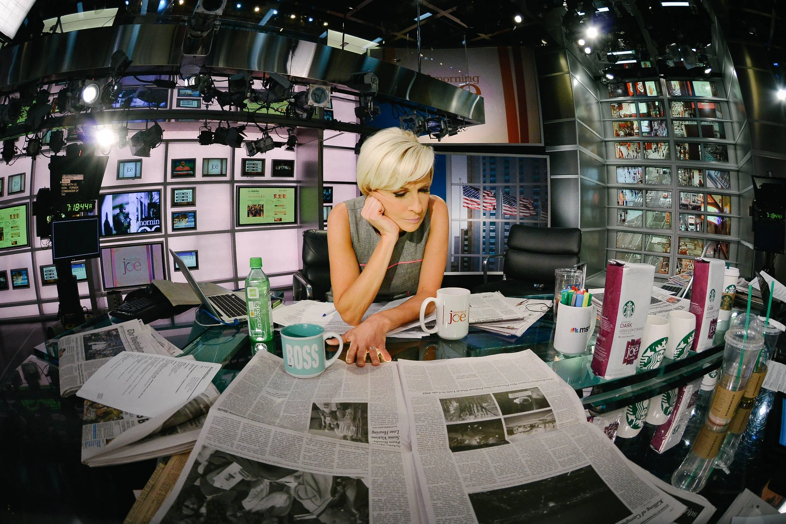 A woman sitting at a cluttered news desk with newspapers, coffee mugs, and various beverages, in a TV studio with multiple screens and American flags in the background.