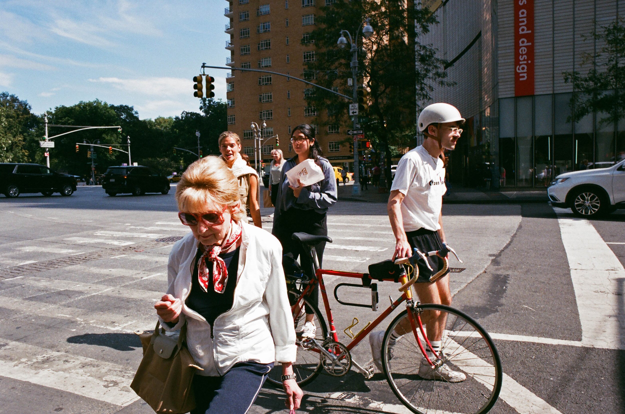 A group of four people crossing the street in an urban area on a sunny day, with cars and buildings in the background. One person is pushing a bicycle.