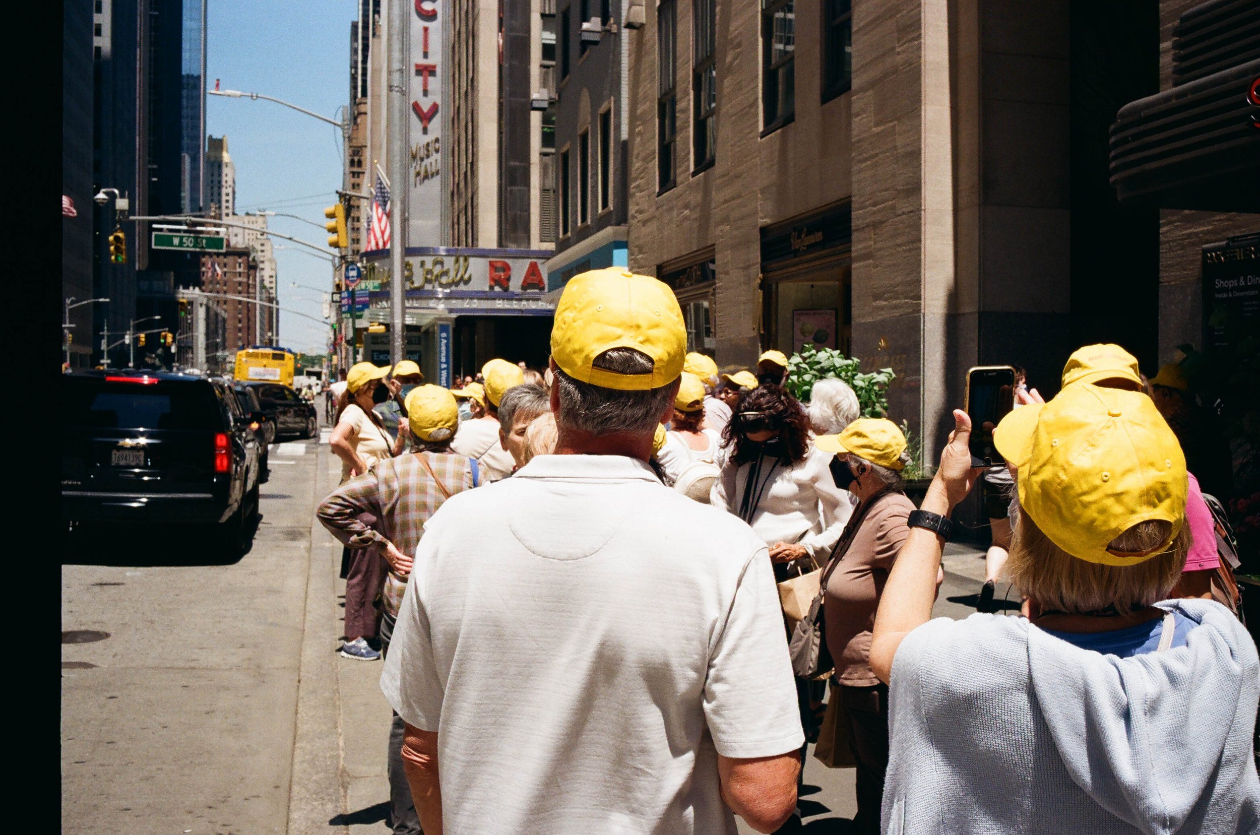 A crowd of people wearing yellow hats standing on a city sidewalk, with some taking photos. Buildings and a street with cars and a yellow bus in the background.