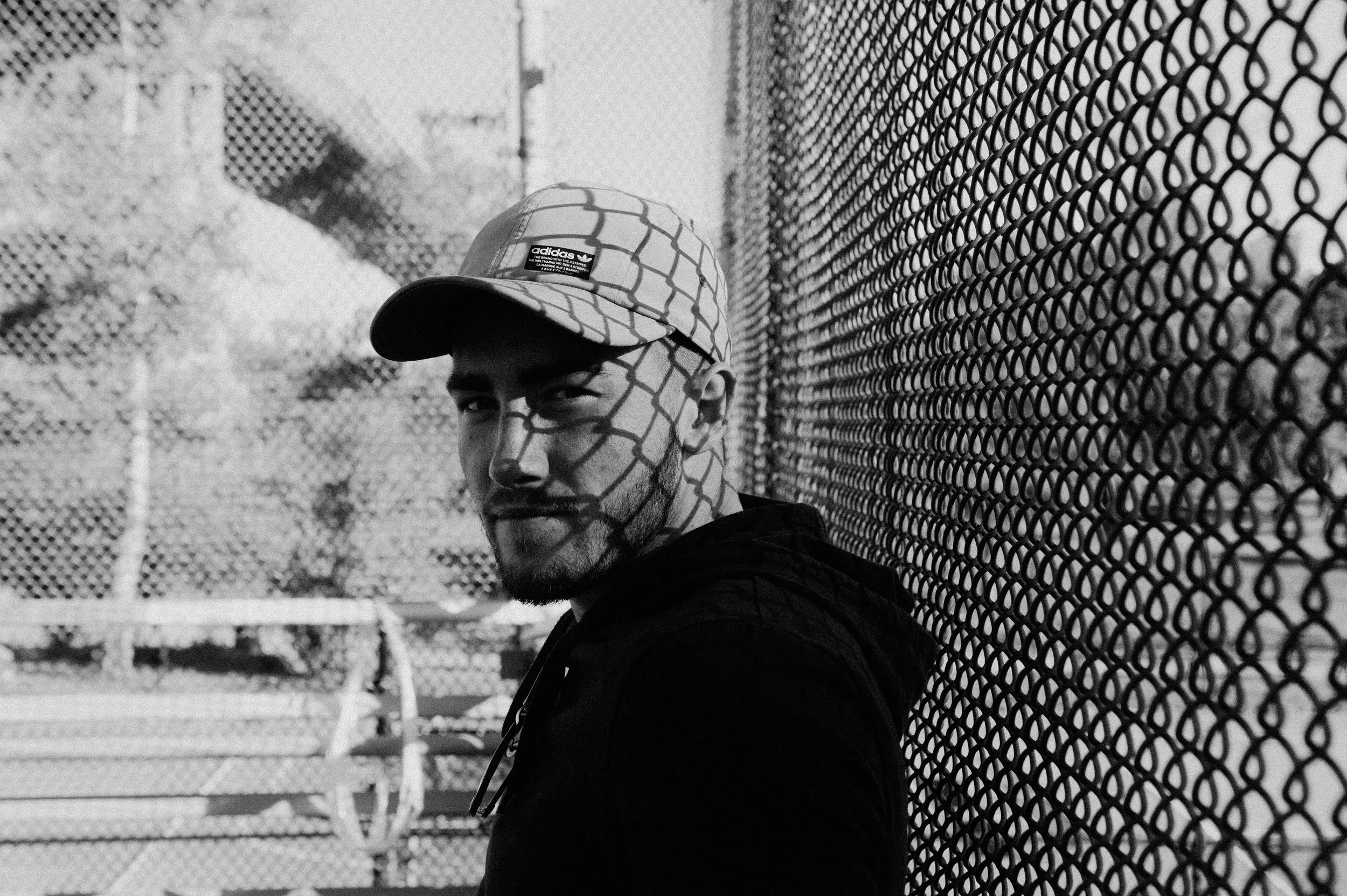 A black and white photo of a man leaning against a chain-link fence, with shadow patterns from the fence on his face. He is wearing a cap and a hoodie.