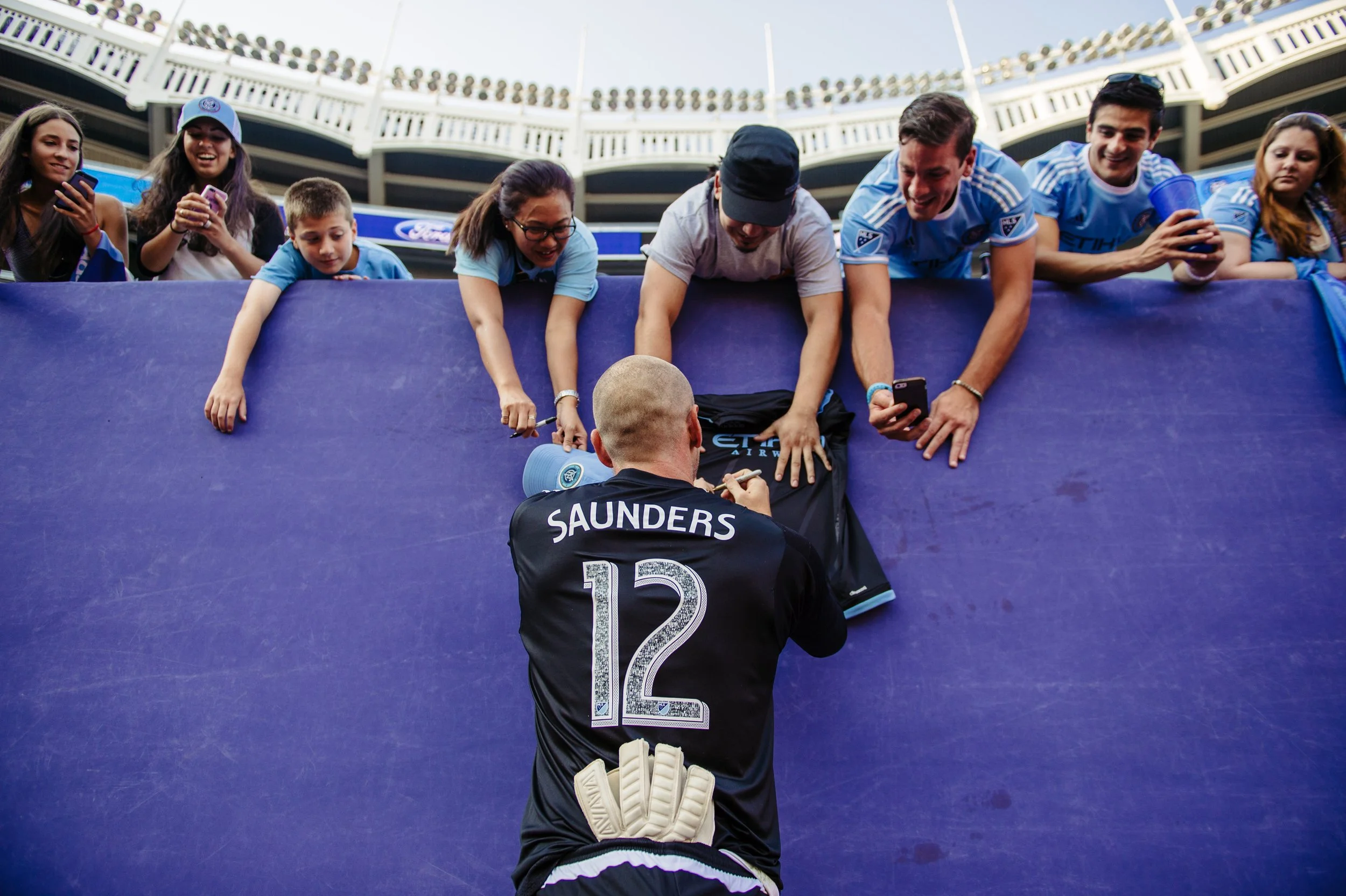 A soccer player in a black jersey with the name Saunders and number 12 signs autographs for excited fans at a stadium, with fans reaching over a barrier to meet him.