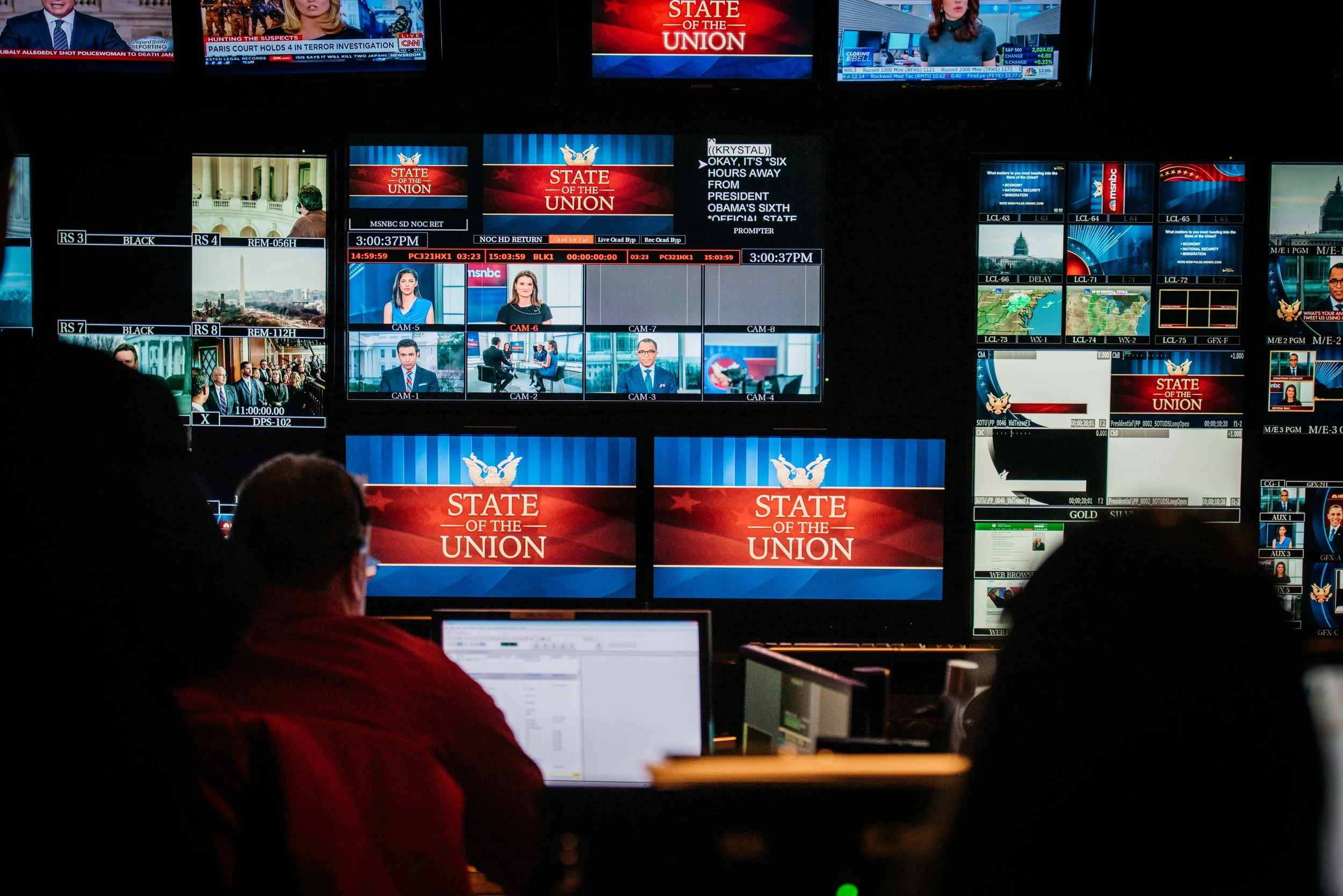 A control room with multiple monitors displaying news coverage and graphics about the State of the Union, with several technicians working at computers.