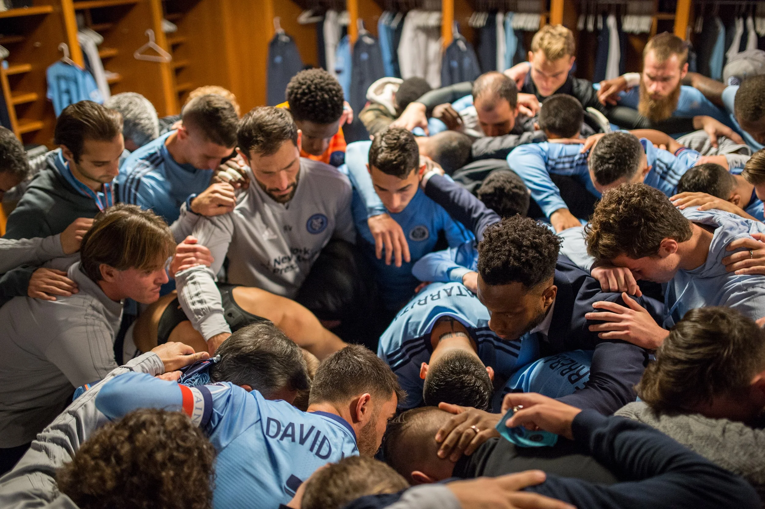 A large group of soccer players in a locker room praying together in a circle with their heads bowed and arms around each other.