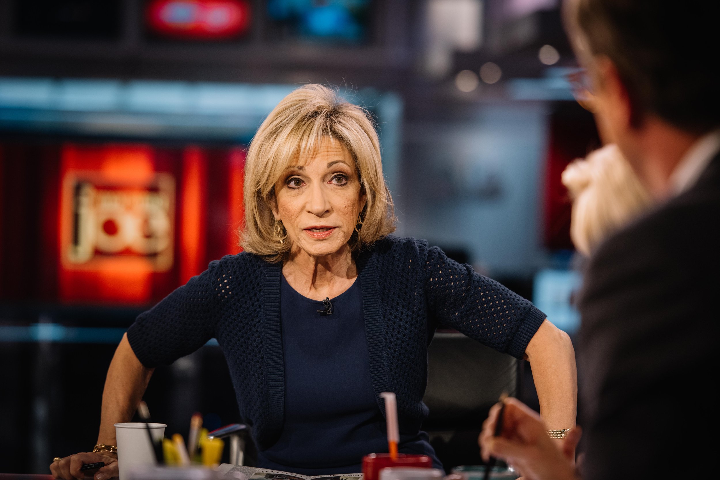 A woman with blonde hair and a blue sweater is talking to a man with dark hair and a suit in a restaurant or cafe setting.