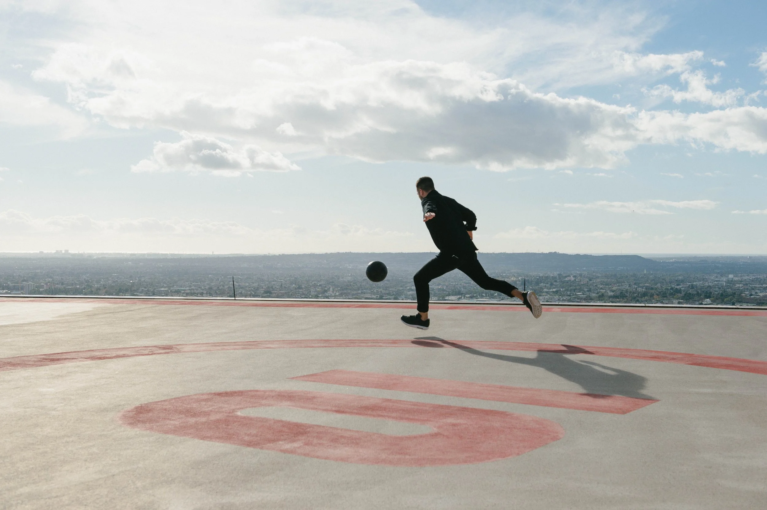 A man in black athletic clothing playing soccer on a rooftop with a view of a city skyline, running after a black soccer ball on a large concrete surface marked with red lines.