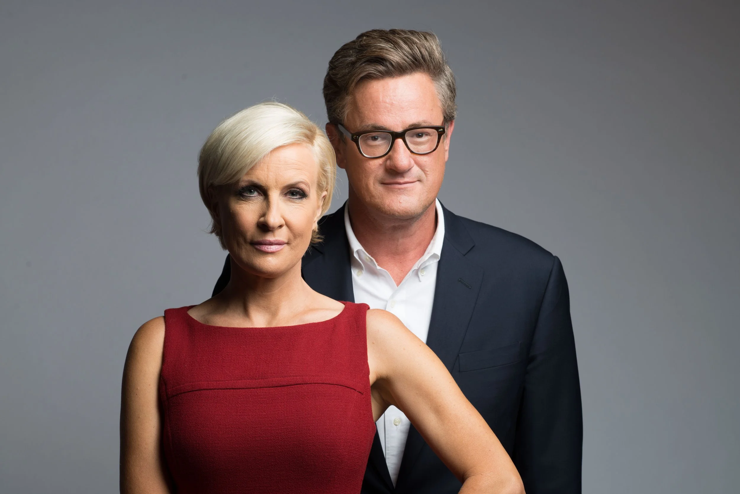 A woman with short blonde hair in a red sleeveless dress and a man with brown hair and glasses in a dark suit and white shirt posing together in front of a gray background.