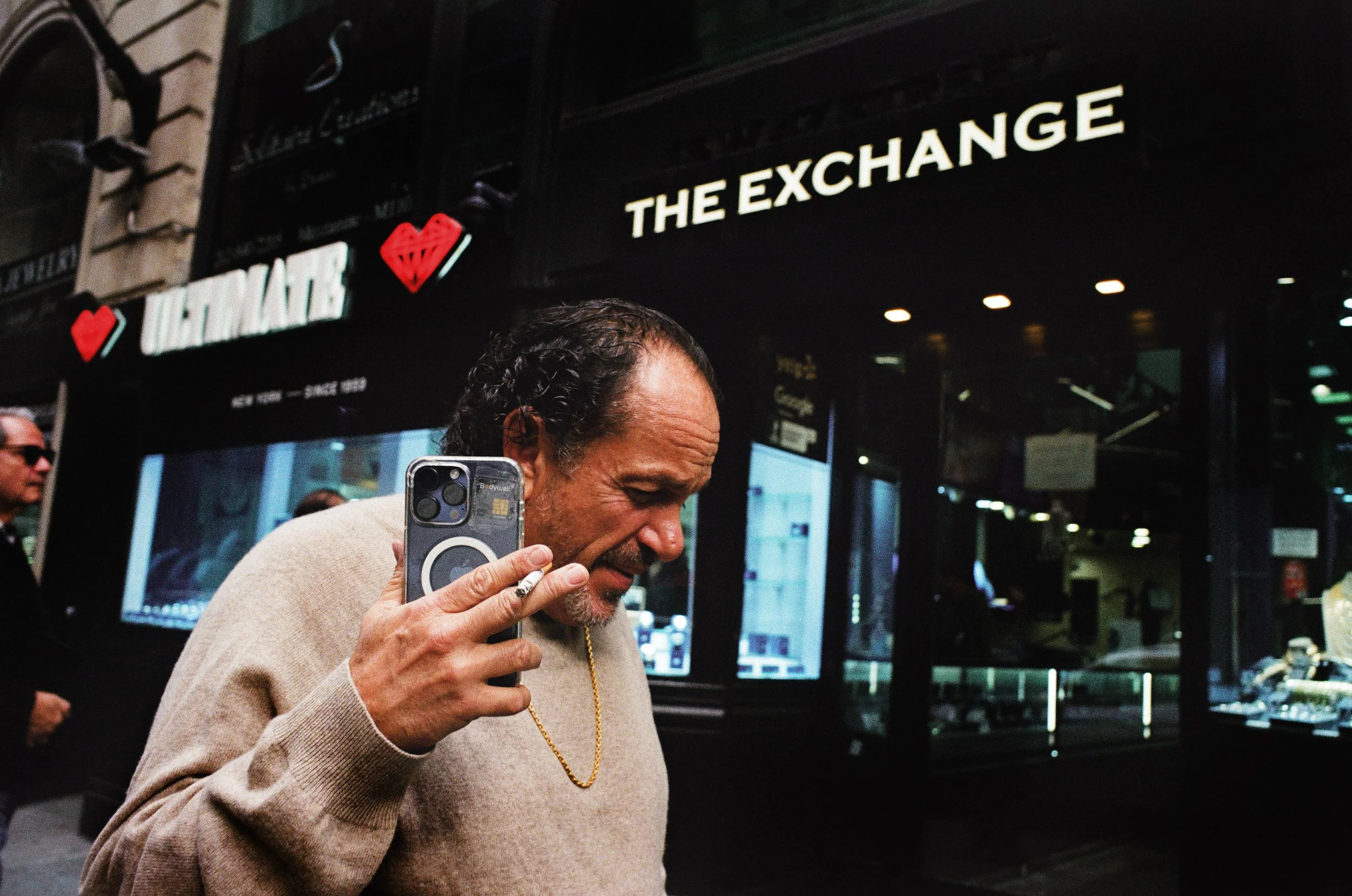 A man with curly hair and a gold chain holding a smartphone with a ring holder, standing in front of a jewelry store called 'The Exchange' with a red heart logo.