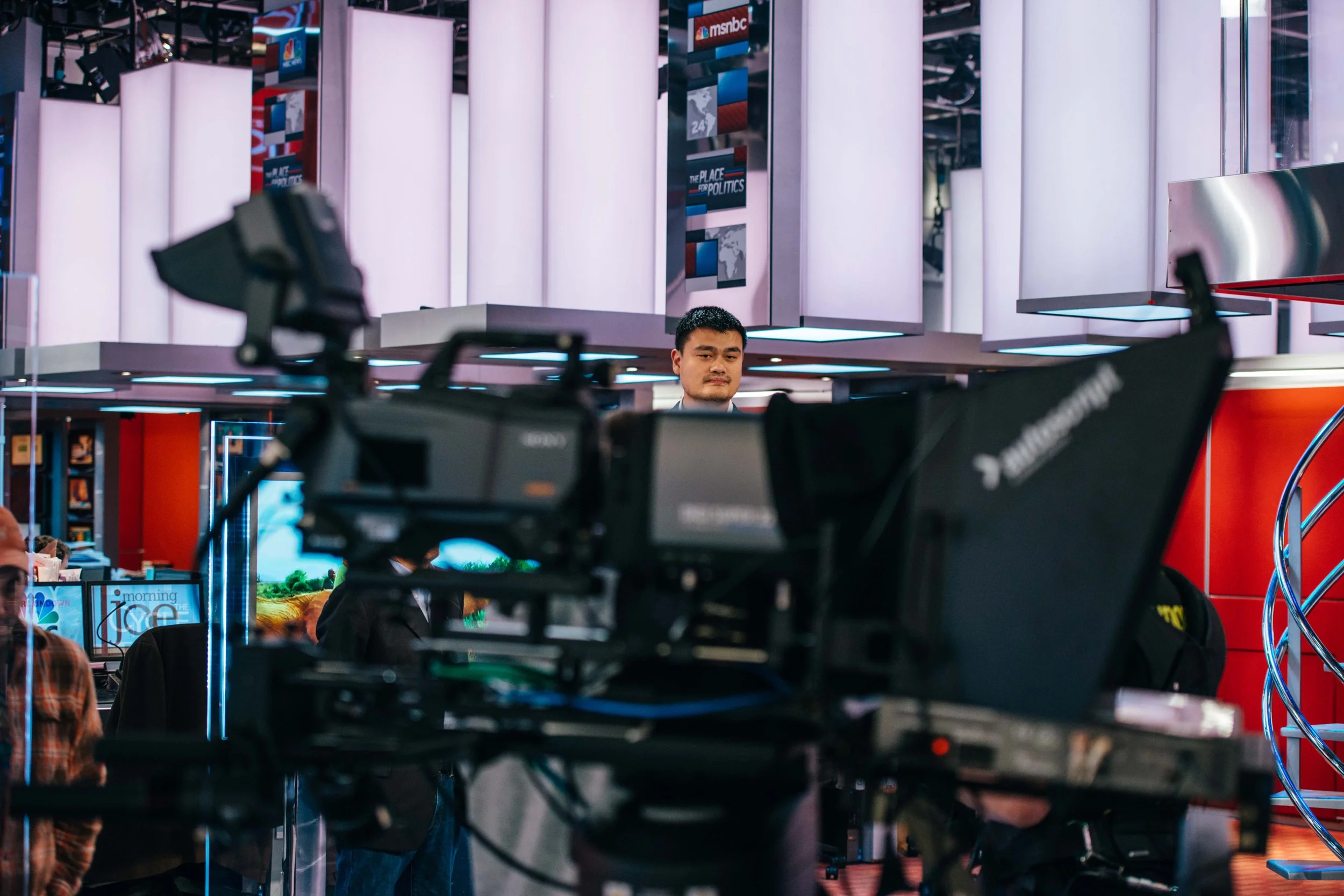 A television news studio with a man standing behind a camera, visible in the background.