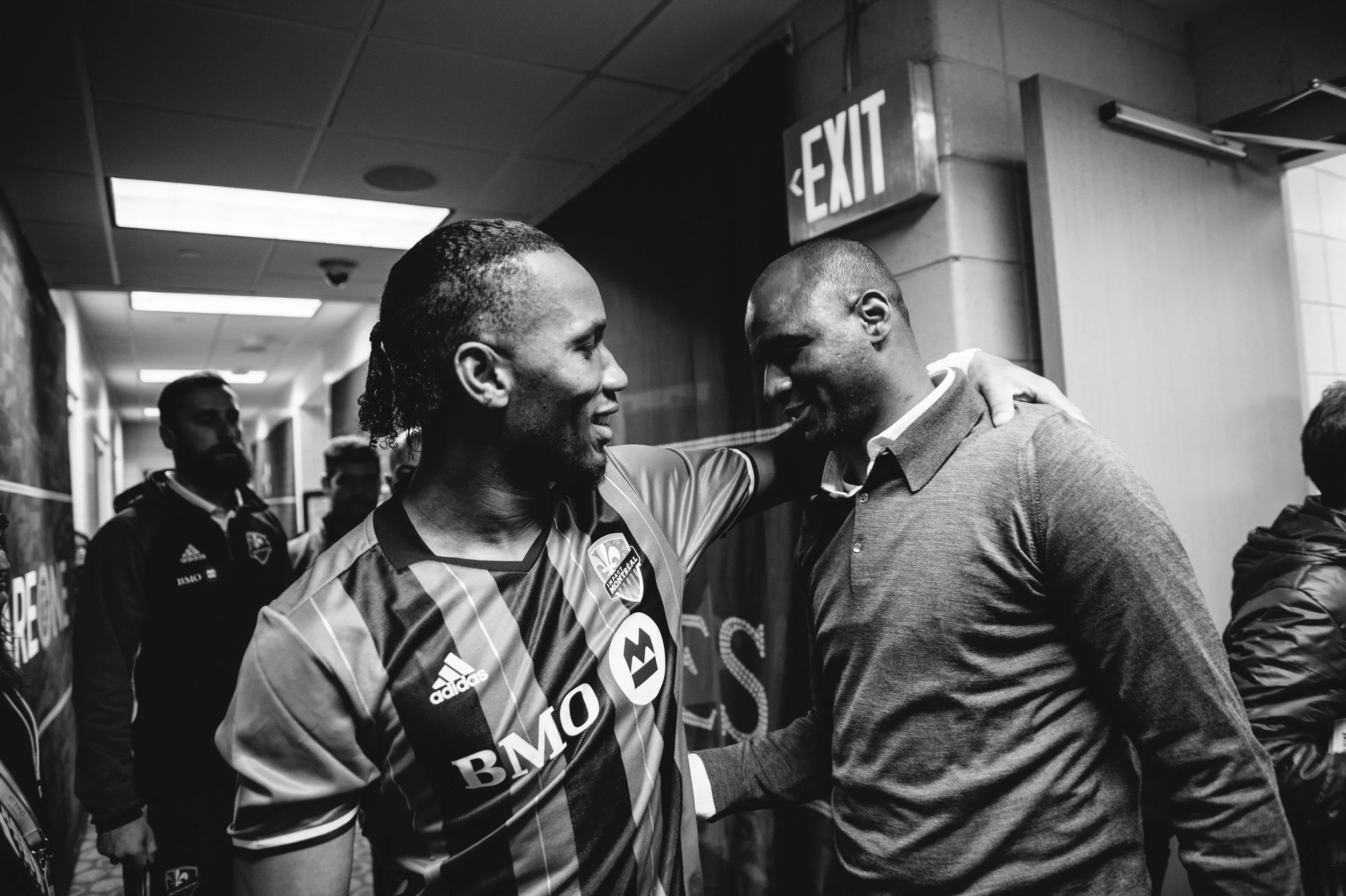 Two men greeting each other with a handshake and friendly gestures in a hallway, one wearing a sports jersey and the other in a button-down shirt.