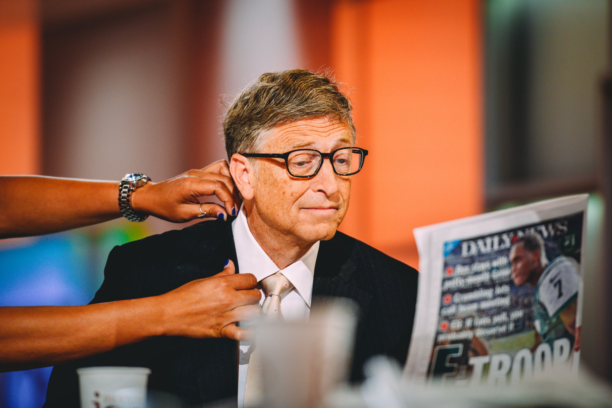A man in glasses and a suit reading a newspaper while being assisted with his necktie or collar by someone else.