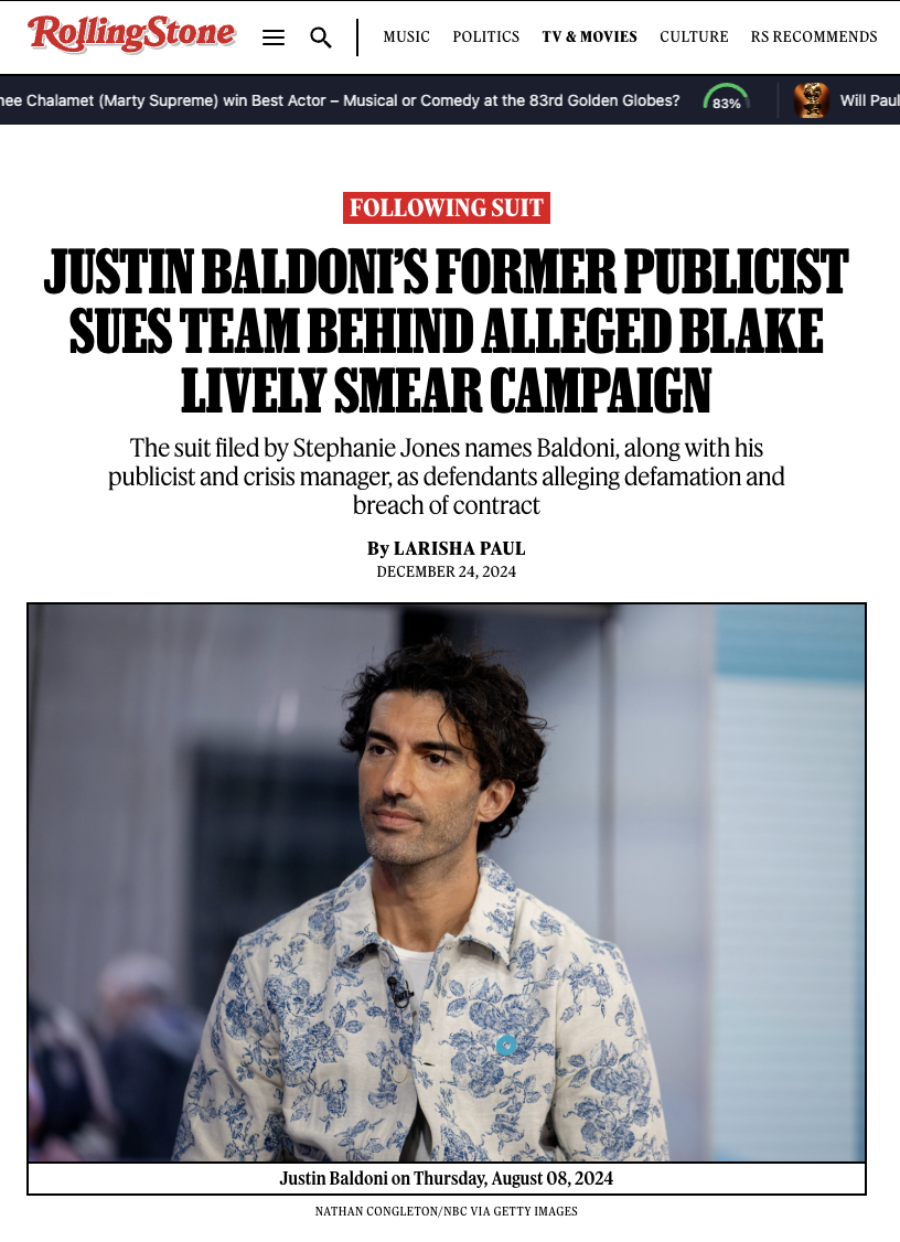 Justin Baldoni with dark, curly hair, wearing a white button-up shirt with blue floral pattern, standing indoors with a neutral facial expression.