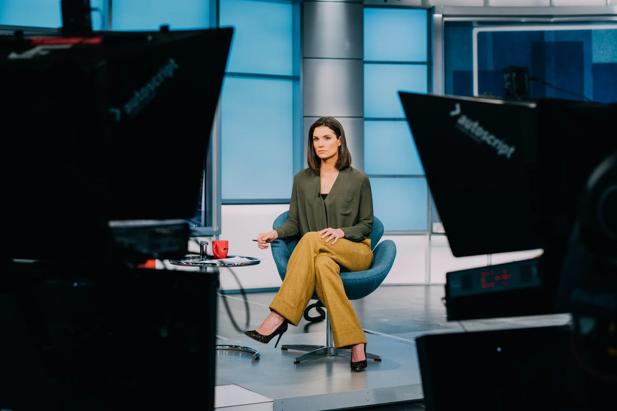 A woman seated in a TV studio or news set, surrounded by cameras and lighting equipment, sitting on a blue chair at a small table with a red mug and some papers.