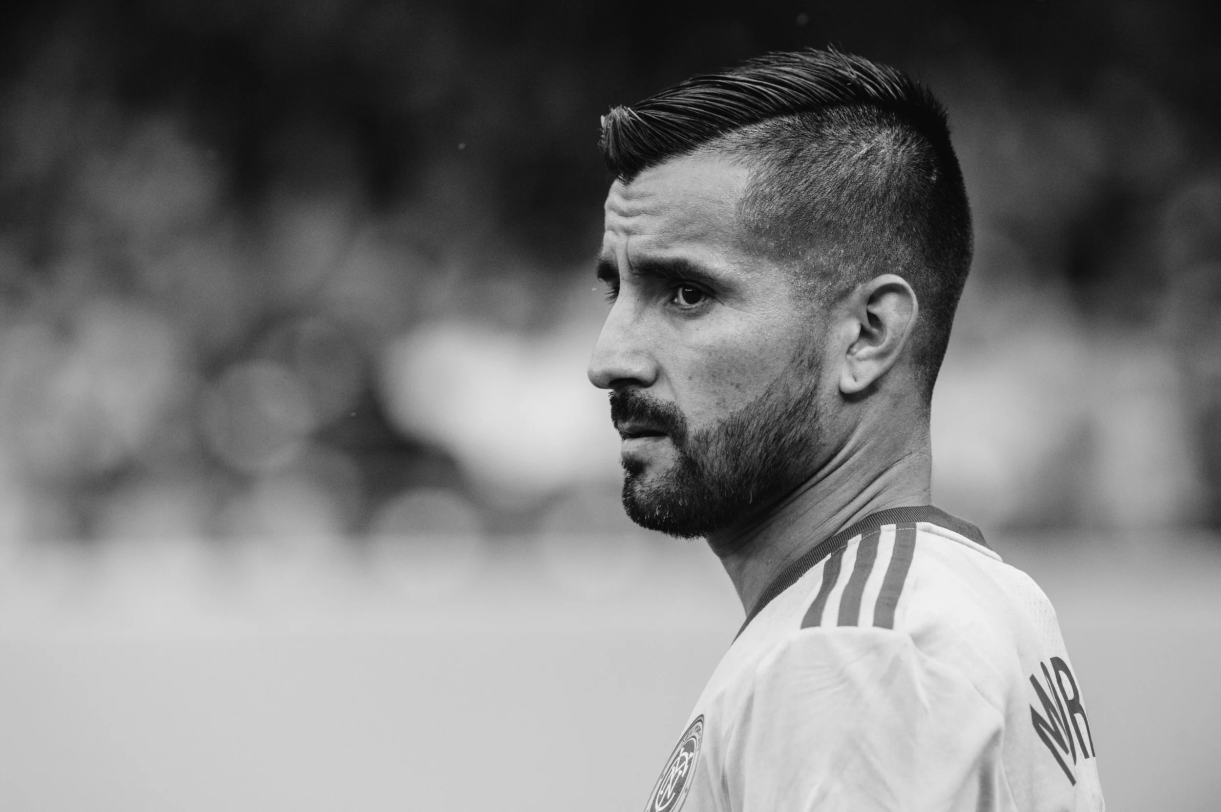 Black and white close-up of a male athlete with a beard, short haircut, wearing a sports jersey, looking to the side with a serious expression.