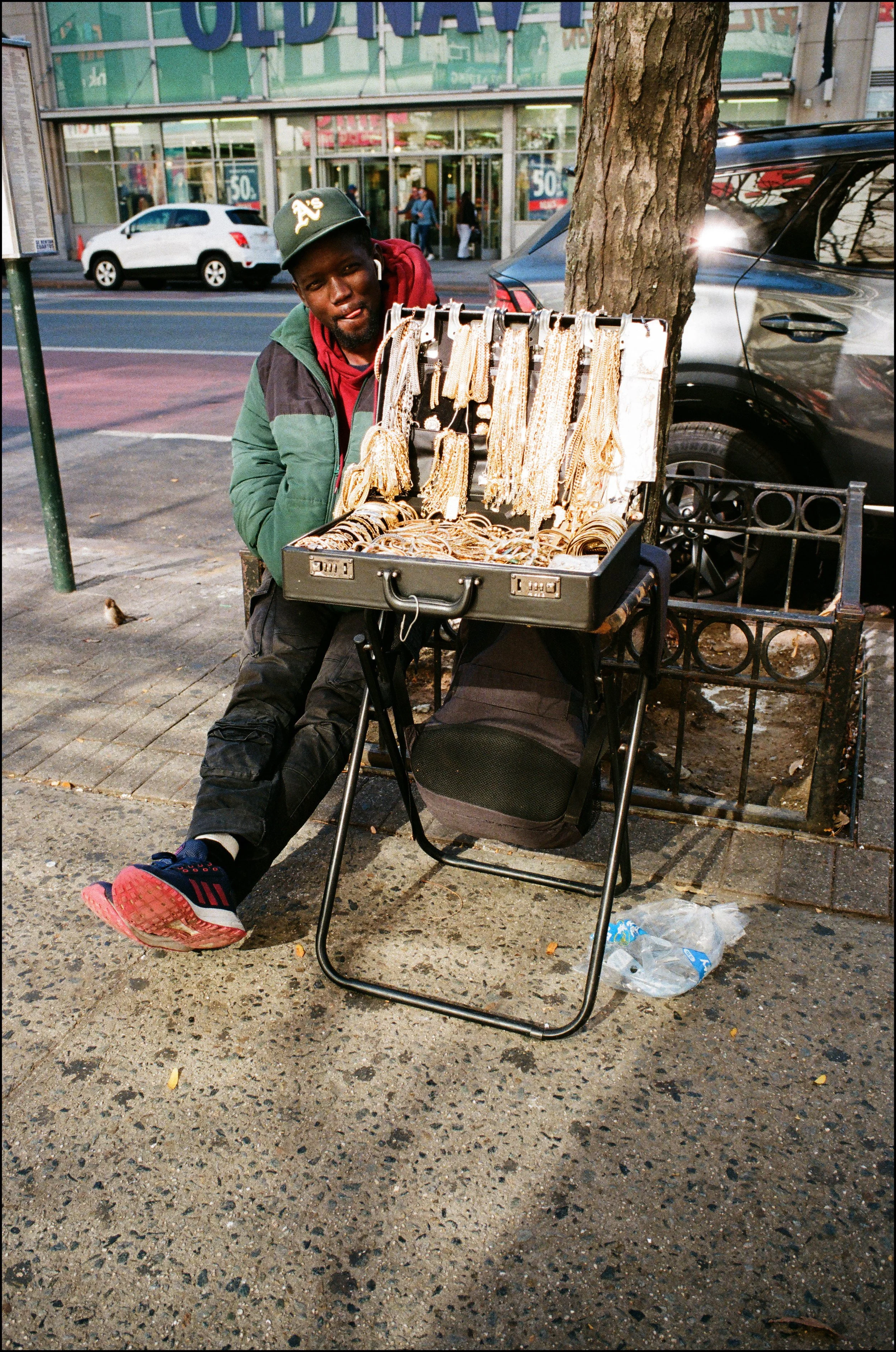 A man sitting on a sidewalk next to a tree displays a suitcase filled with gold jewelry, including necklaces, bracelets, and rings, for sale.