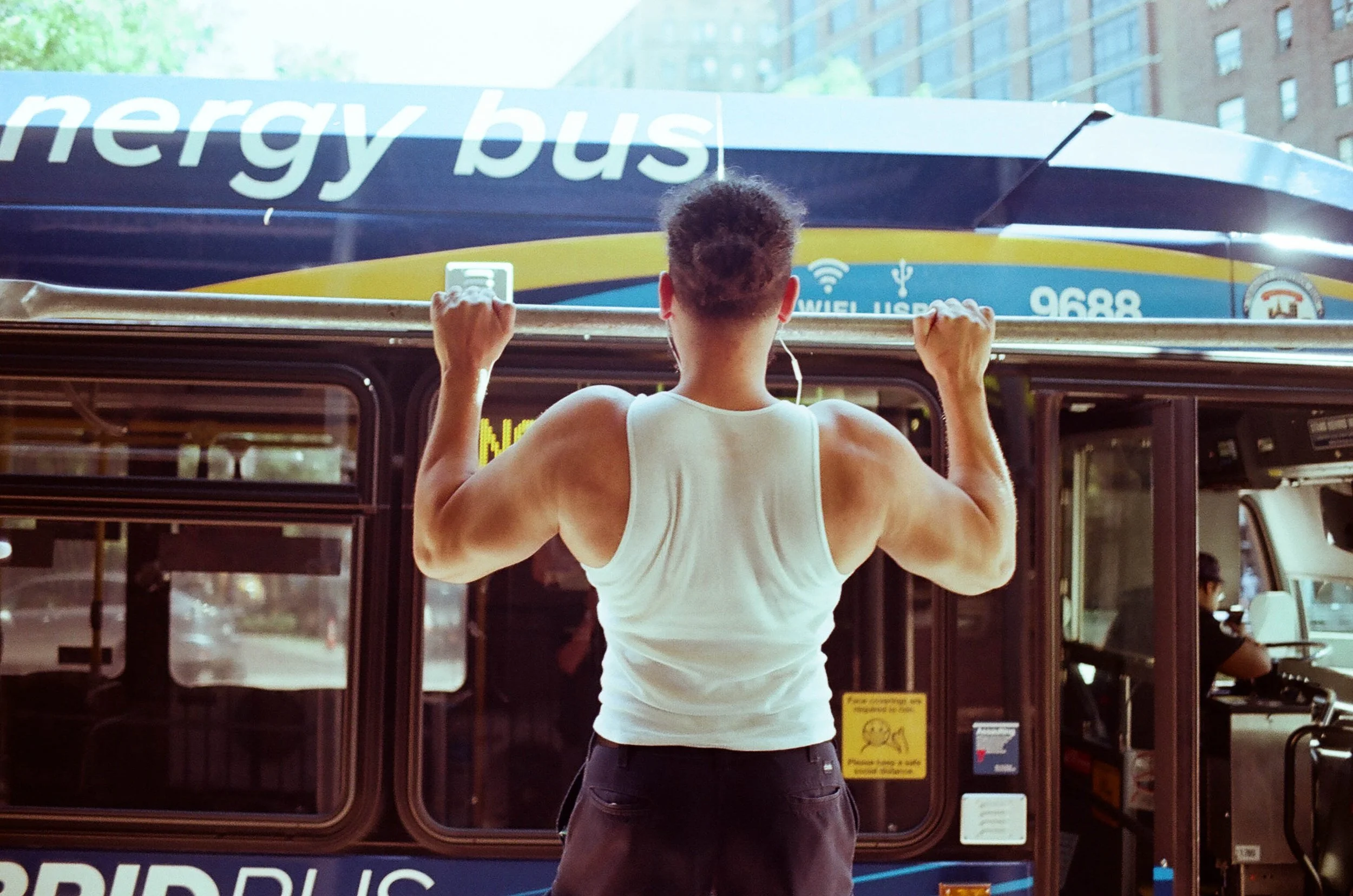 A muscular man in a white tank top does pull-ups on a city bus, with an energy bus visible in the background.