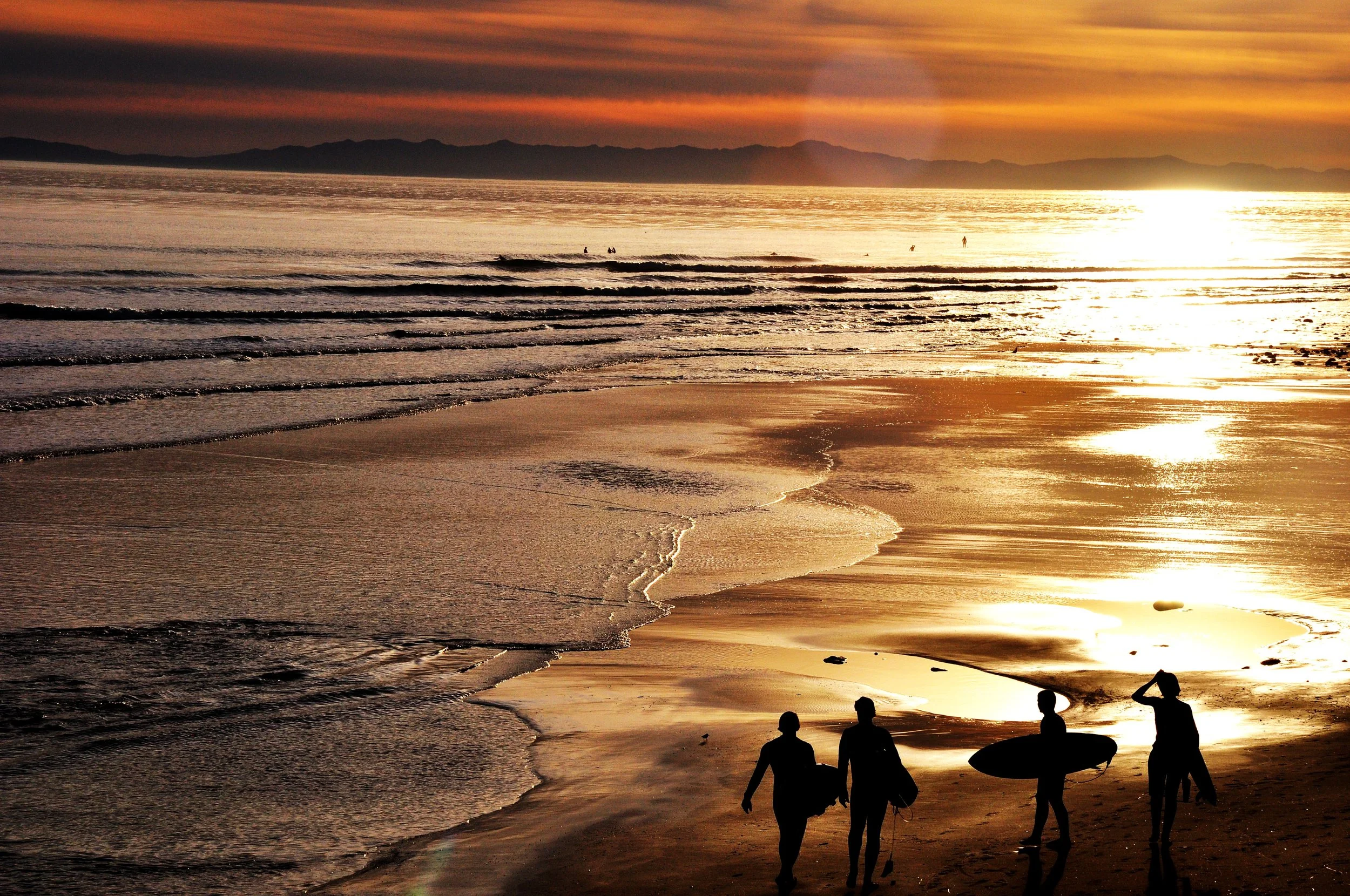 Silhouettes of four people walking on the beach during sunset, with one carrying a surfboard, and the ocean waves approaching the shore.