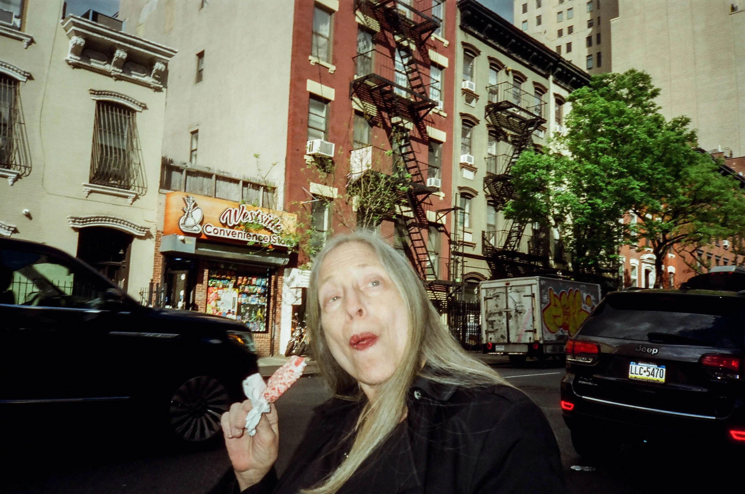 A woman with long gray hair standing on a city street, holding a food item in her hand. Behind her, there are parked cars, a convenience store sign, trees, and residential buildings with fire escapes.