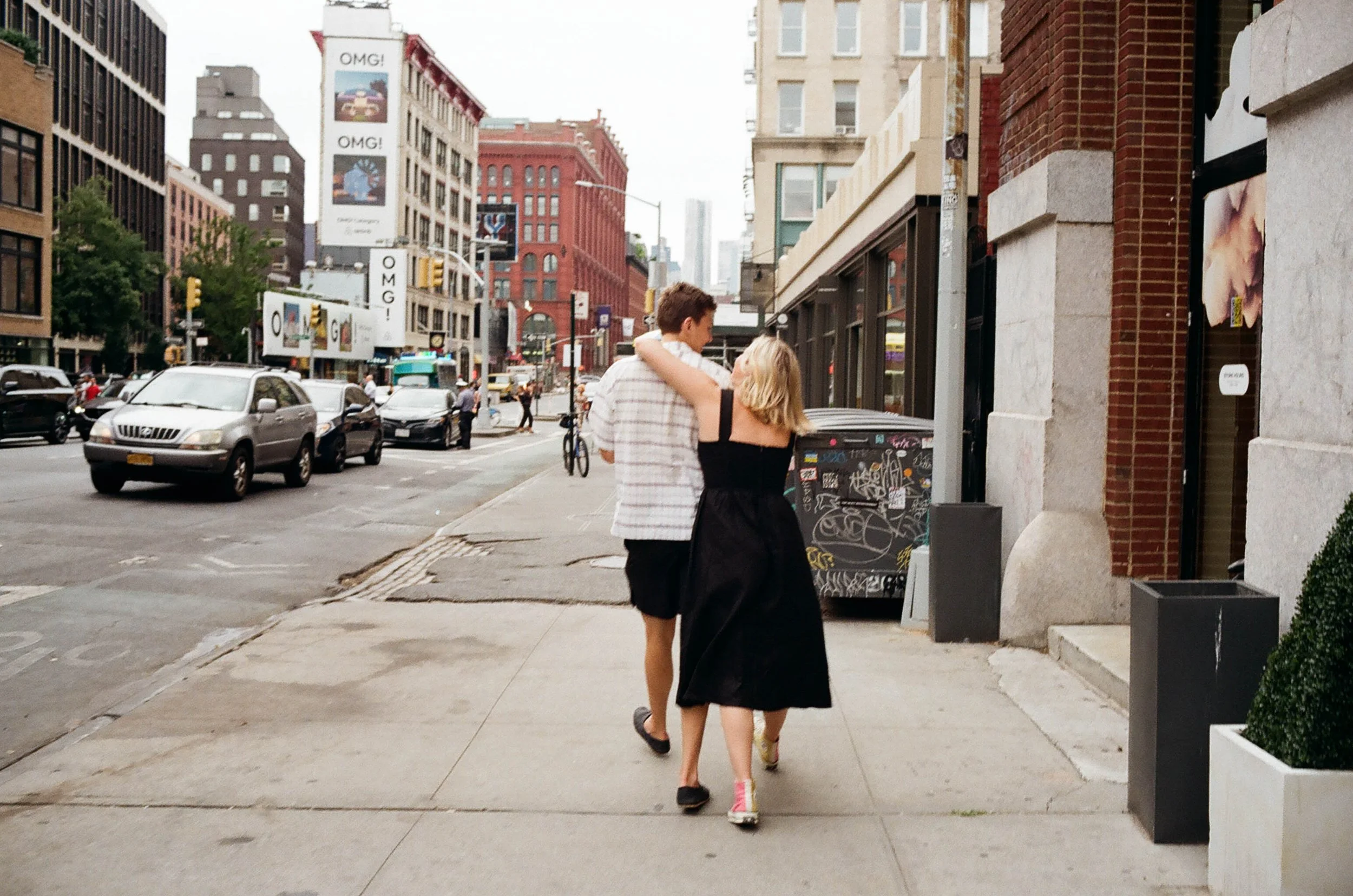 A young couple walking on a city sidewalk, with the man wearing a striped shirt and shorts, and the woman in a black dress and sneakers, with their arms around each other.