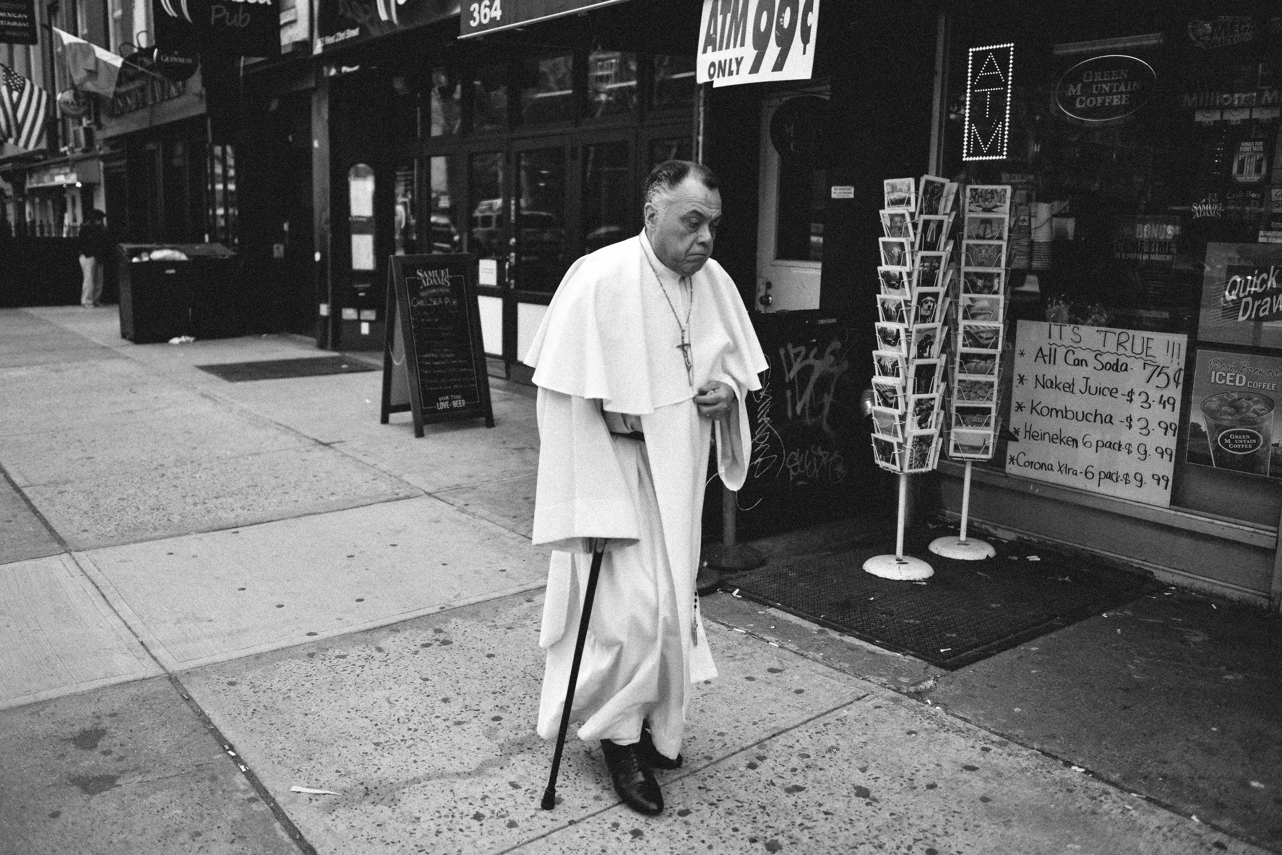 A man dressed as Pope Francis walking on a city sidewalk, holding a cane, outside a shop with signs and advertisements.