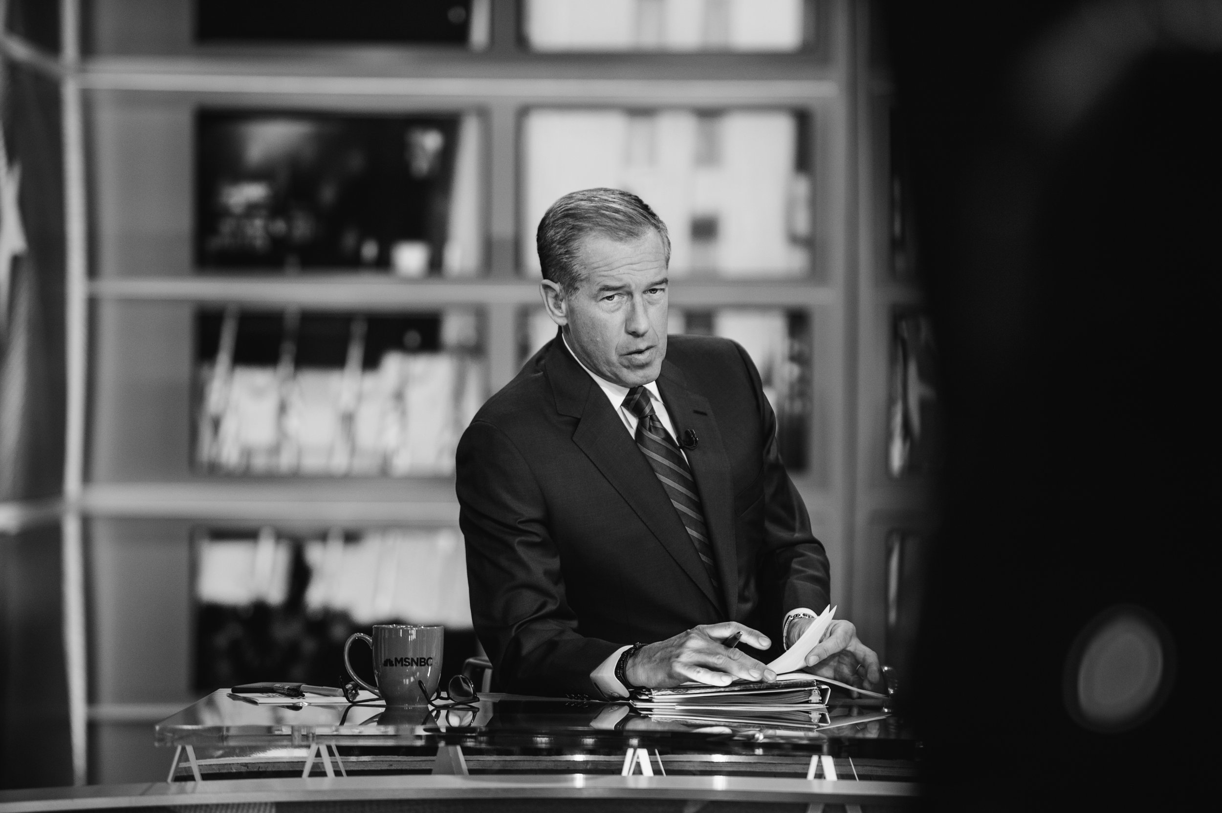 Black and white photo of a man in a suit sitting at a desk with papers, a mug, and glasses, on a TV show set with bookshelves in the background.