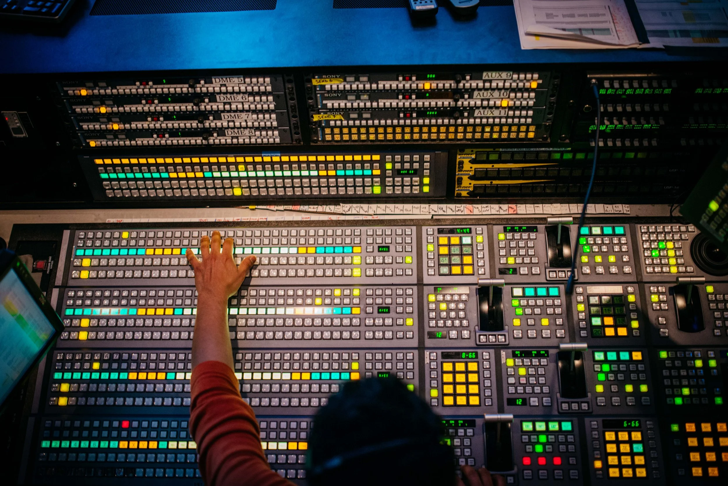 A person operating a professional audio mixing console with numerous colorful controls, screens, and equipment, viewed from above in a studio or control room.