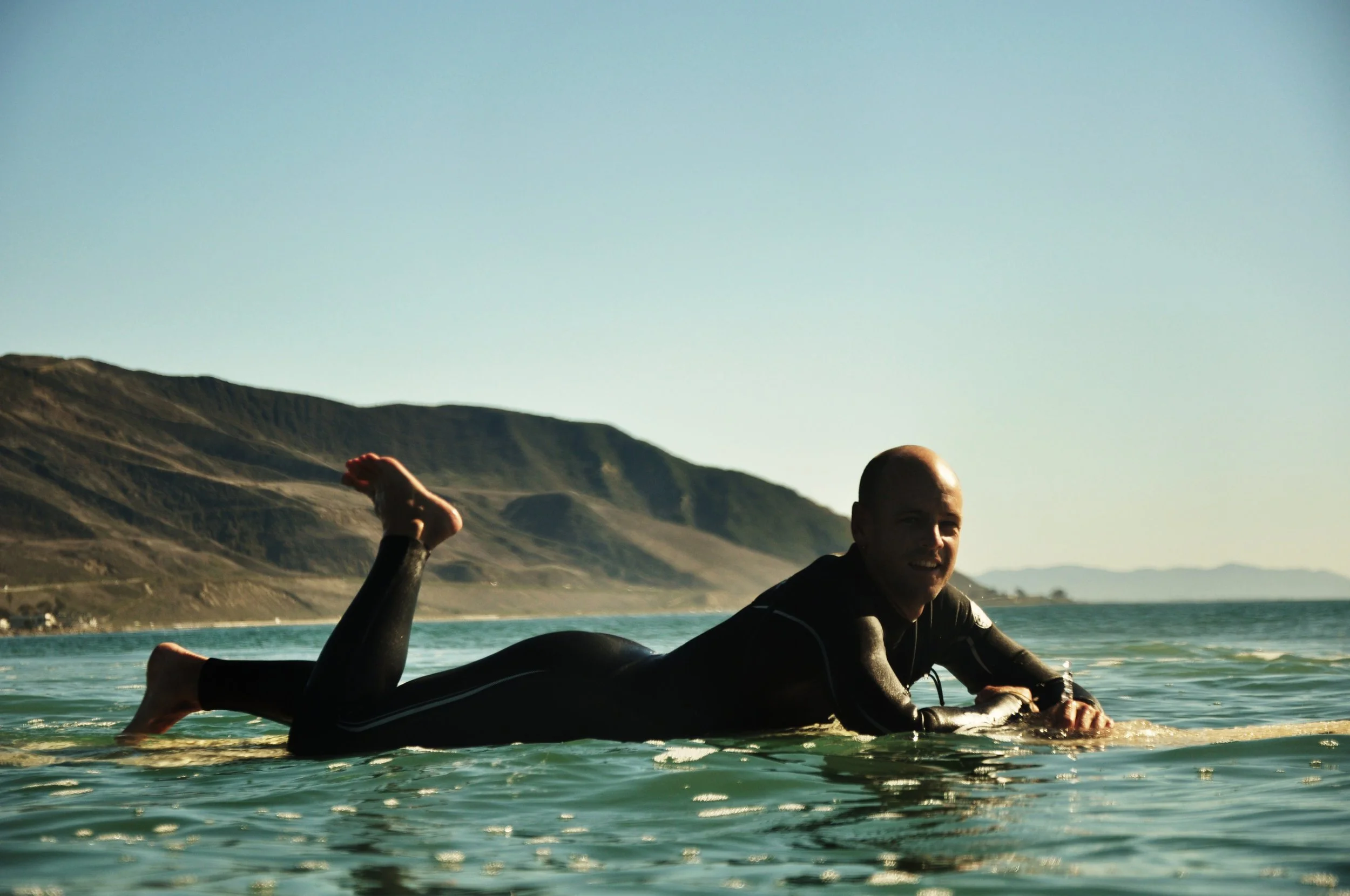 A man in a wetsuit lying on a surfboard in the water with a mountainous coastline in the background.