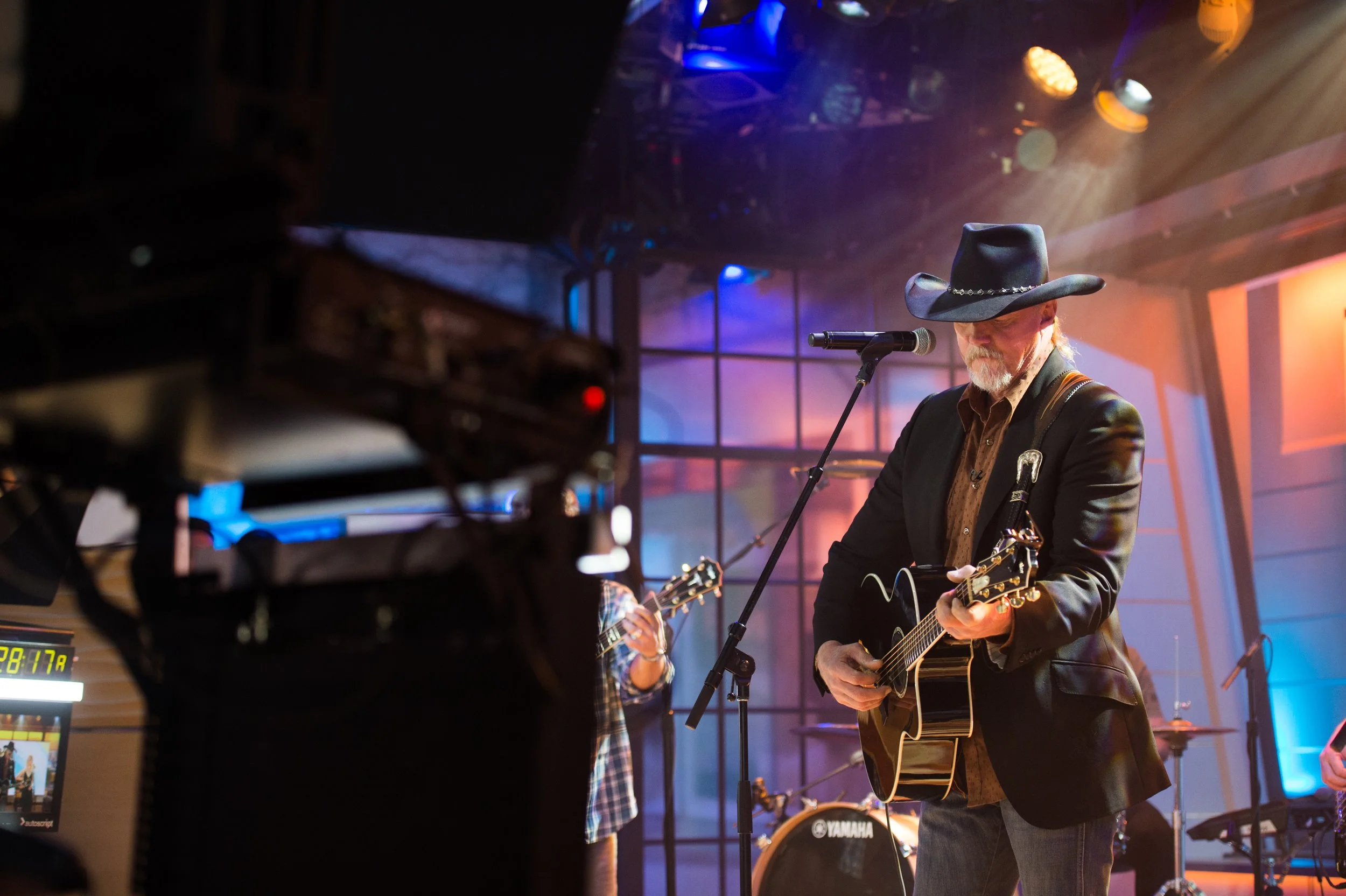 A man wearing a cowboy hat and blazer playing an acoustic guitar on stage with colorful lighting.