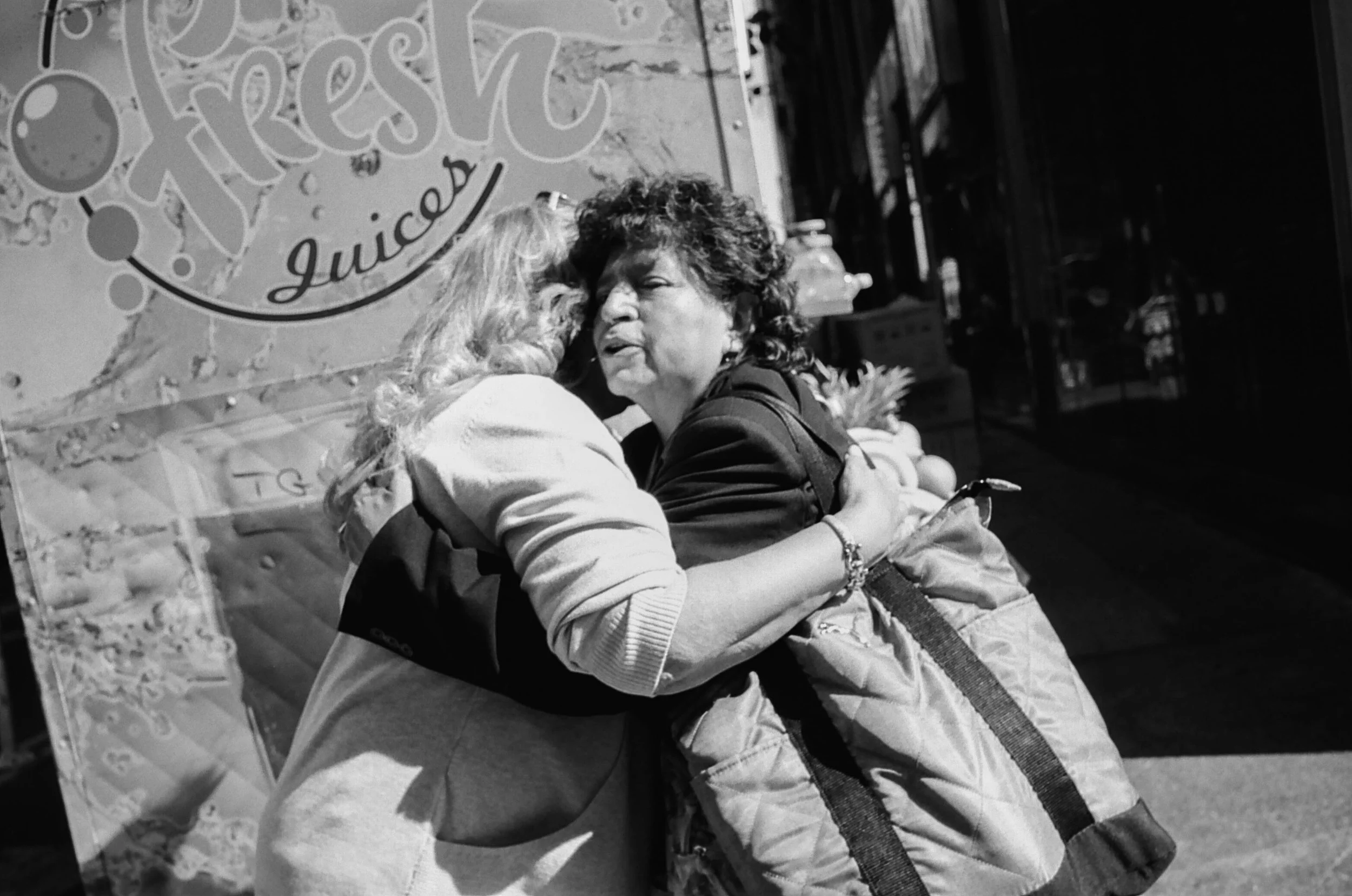 Two women hugging on the sidewalk in front of a sign for Fresh Juices.