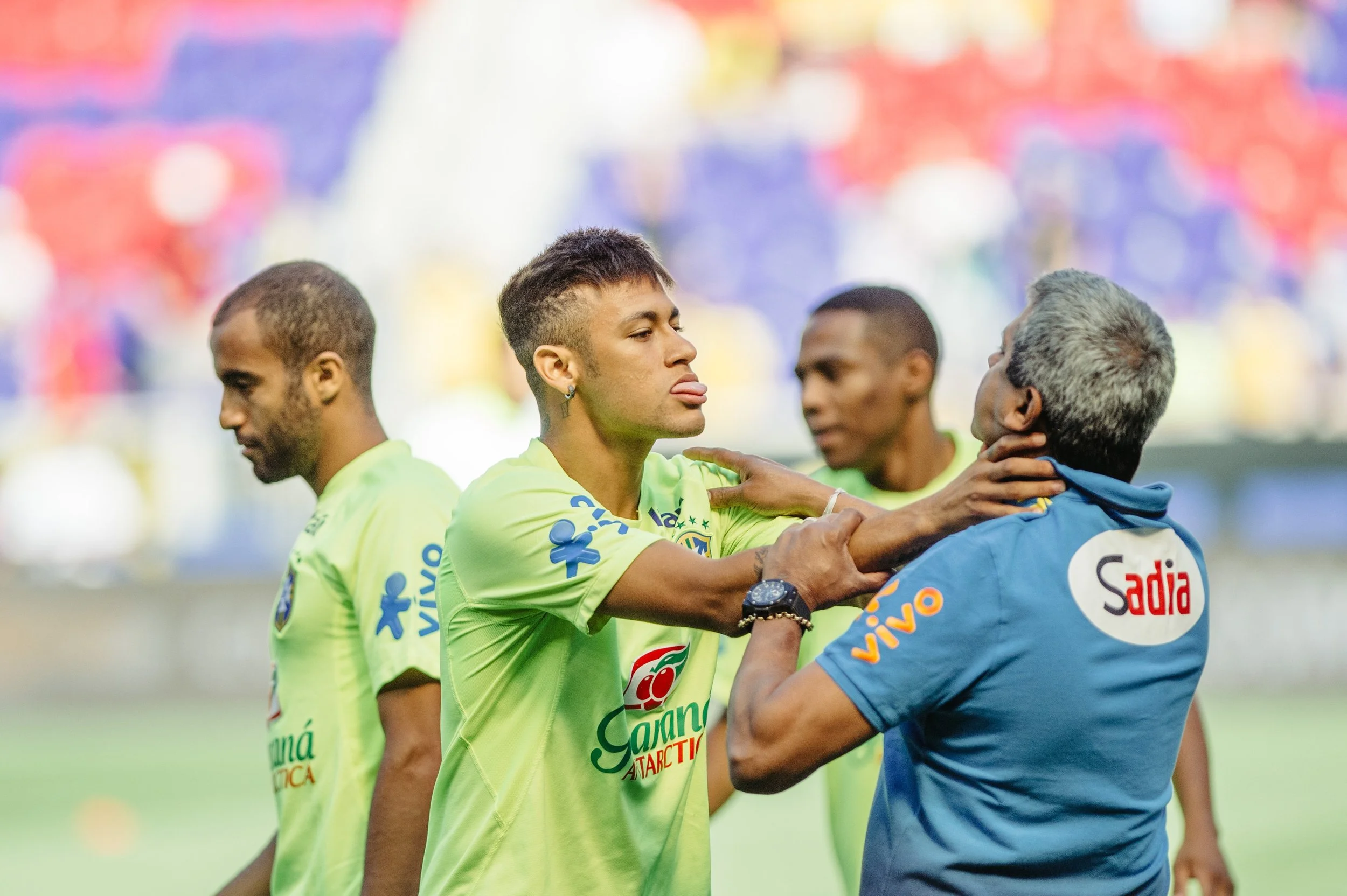A soccer player in a green jersey is being held back by a man in a blue shirt, while three other players in green jerseys look on. The scene appears tense, possibly after a match.