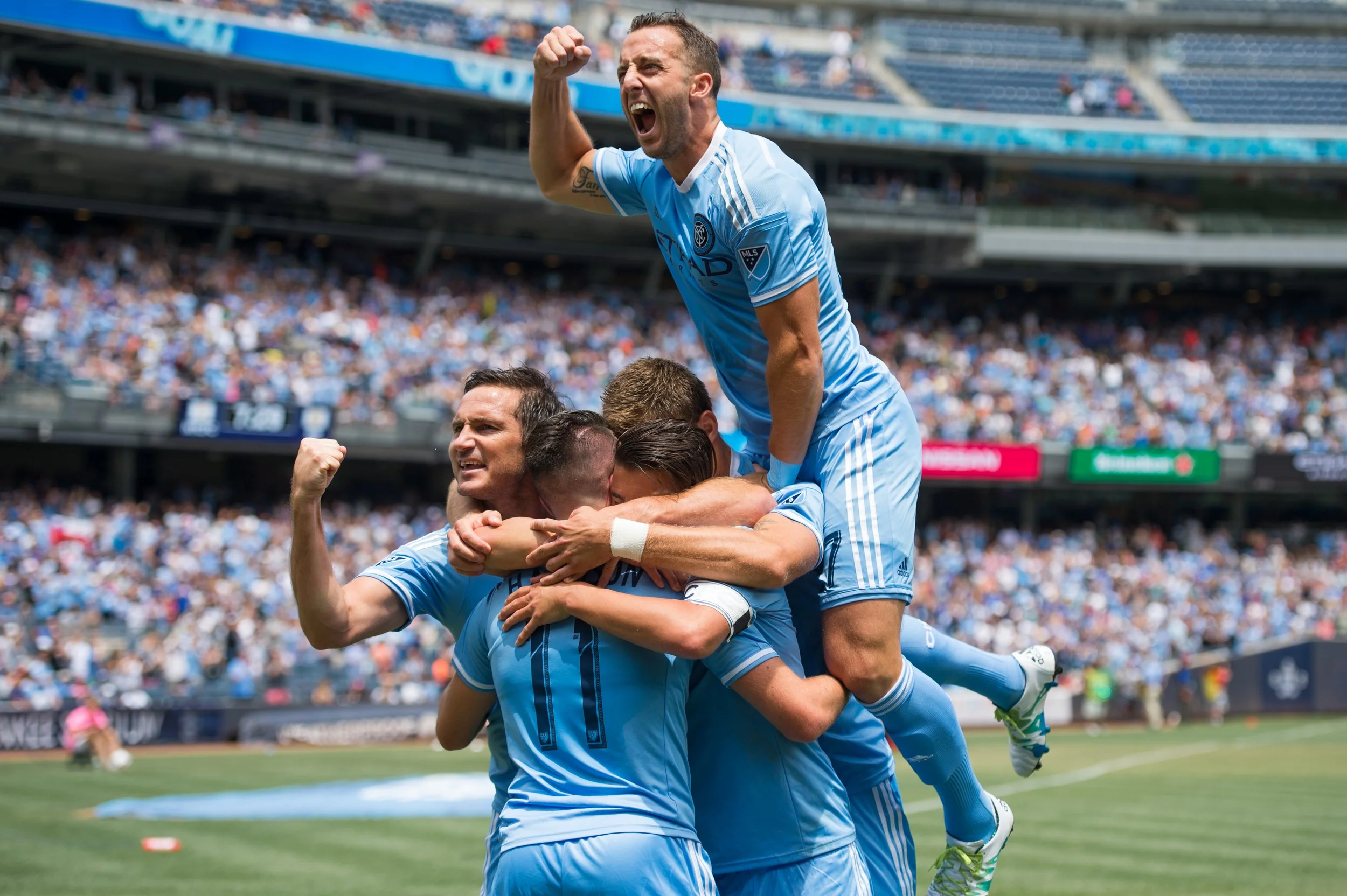A group of soccer players in light blue jerseys celebrate a goal on the field, with one player on top raising his fist in victory, while others embrace in a group hug, with a large crowd in the stadium background.