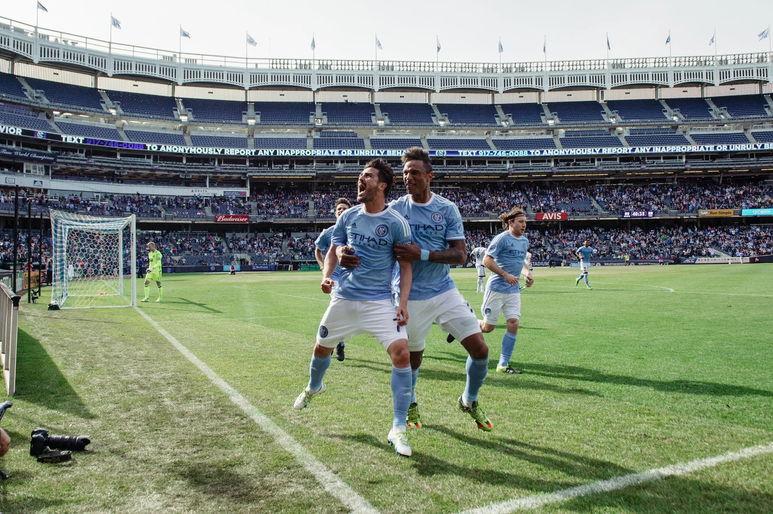 Soccer players in blue jerseys celebrating on the field of a stadium after scoring a goal, with fans in the stands.