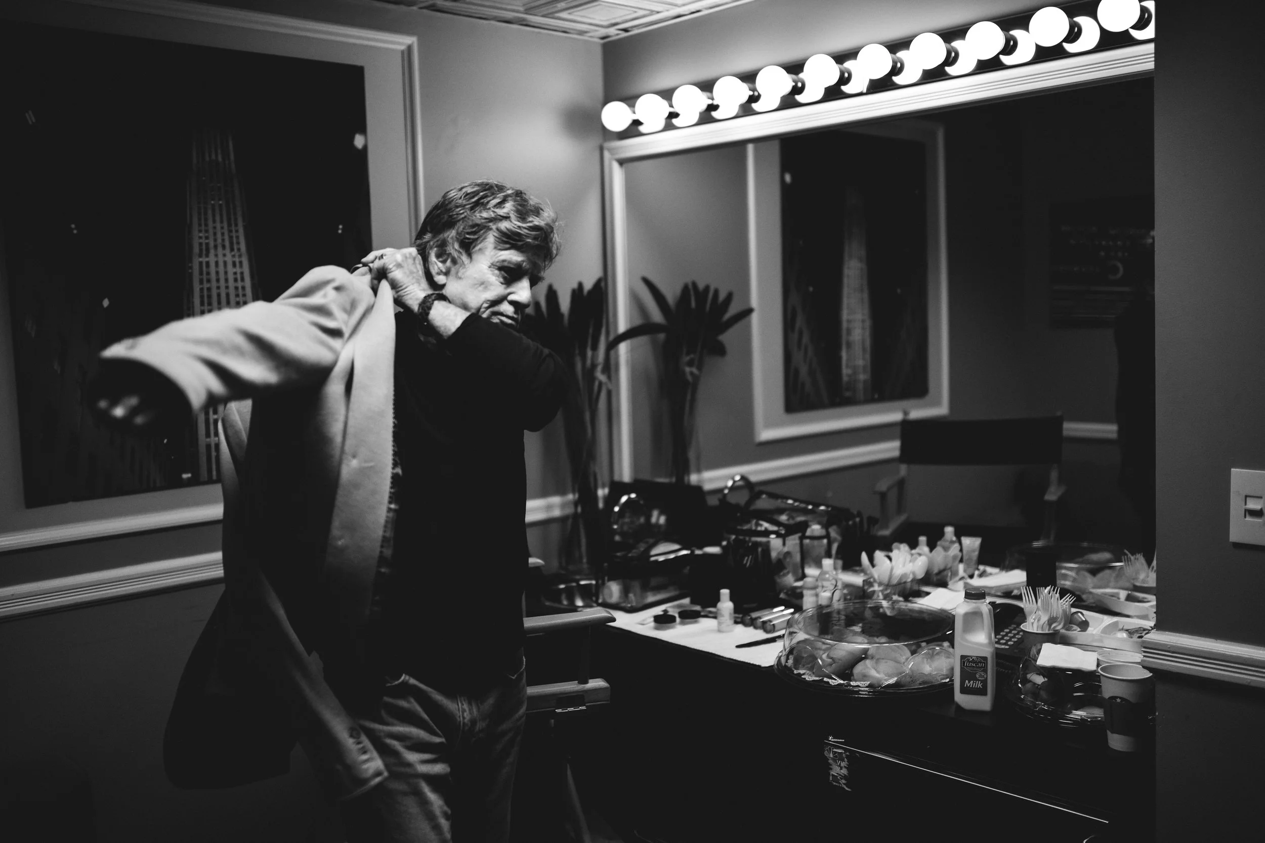 Man putting on a jacket in a dressing room with makeup and styling supplies on a table.