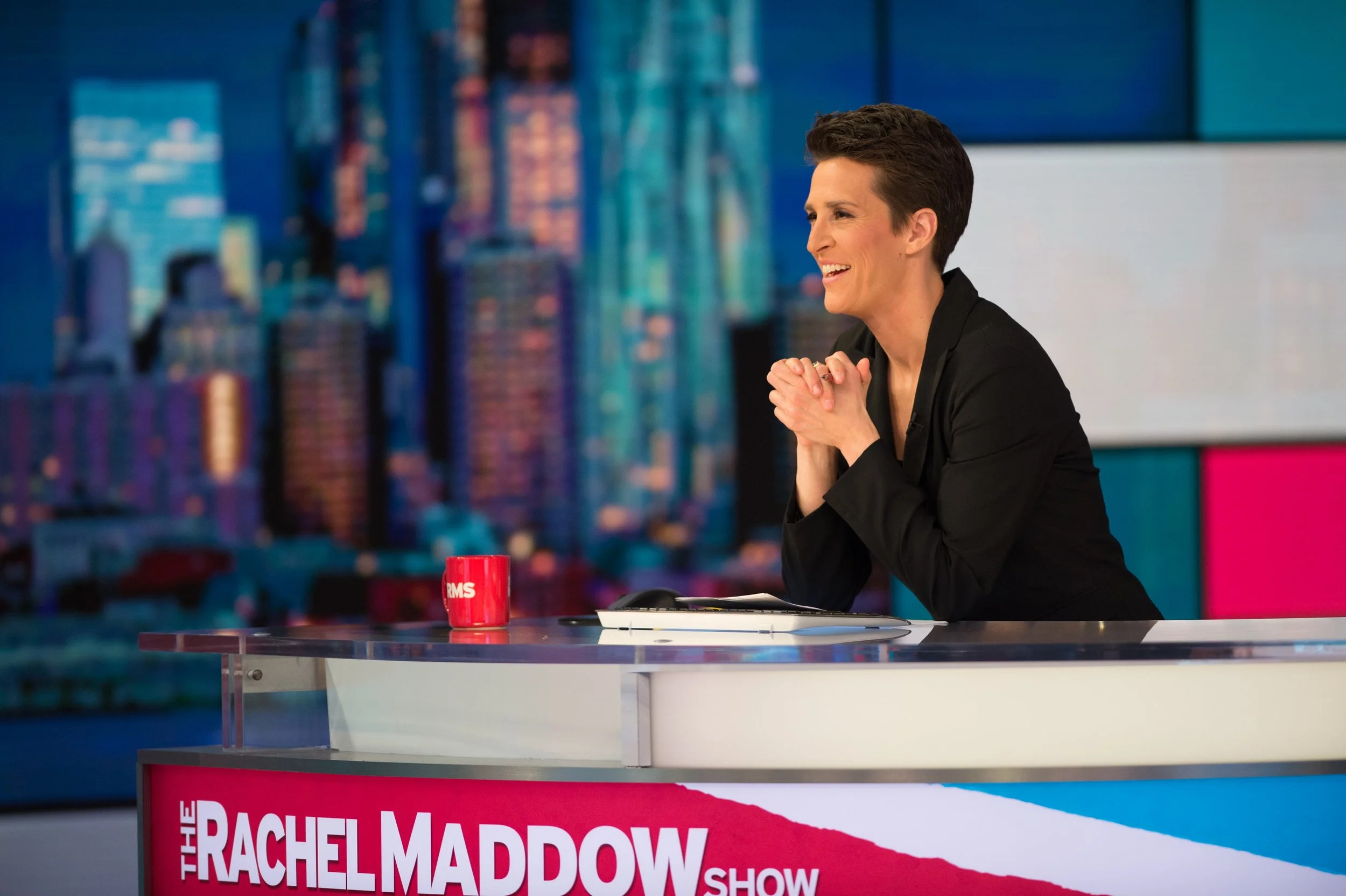 Rachel Maddow host on set of 'The Rachel Maddow Show,' sitting at a desk with a cityscape background, smiling with hands clasped.