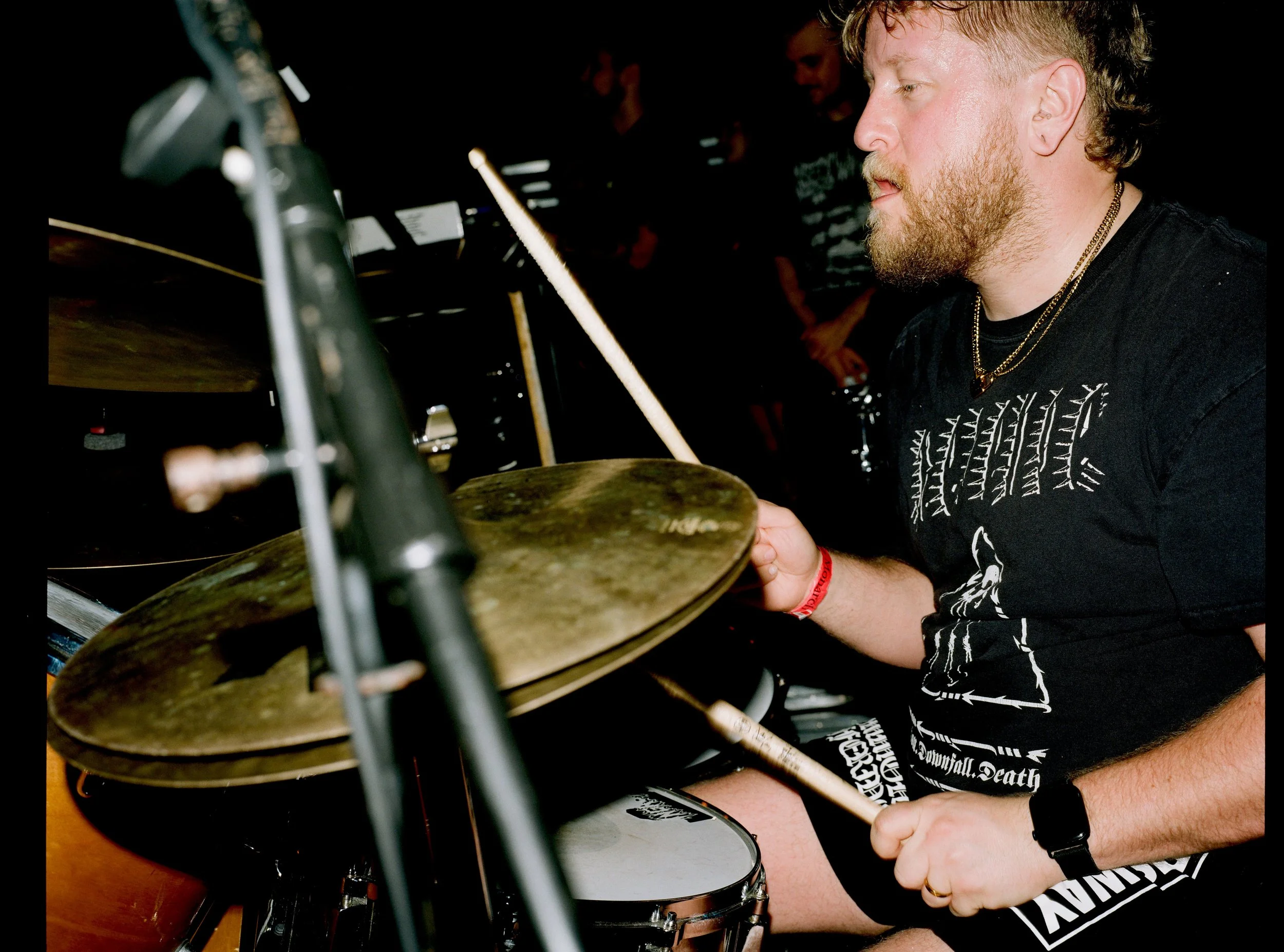 A man with a beard and short hair playing drums with a focused expression in a dark room, wearing a black t-shirt with white graphics and gold necklaces.