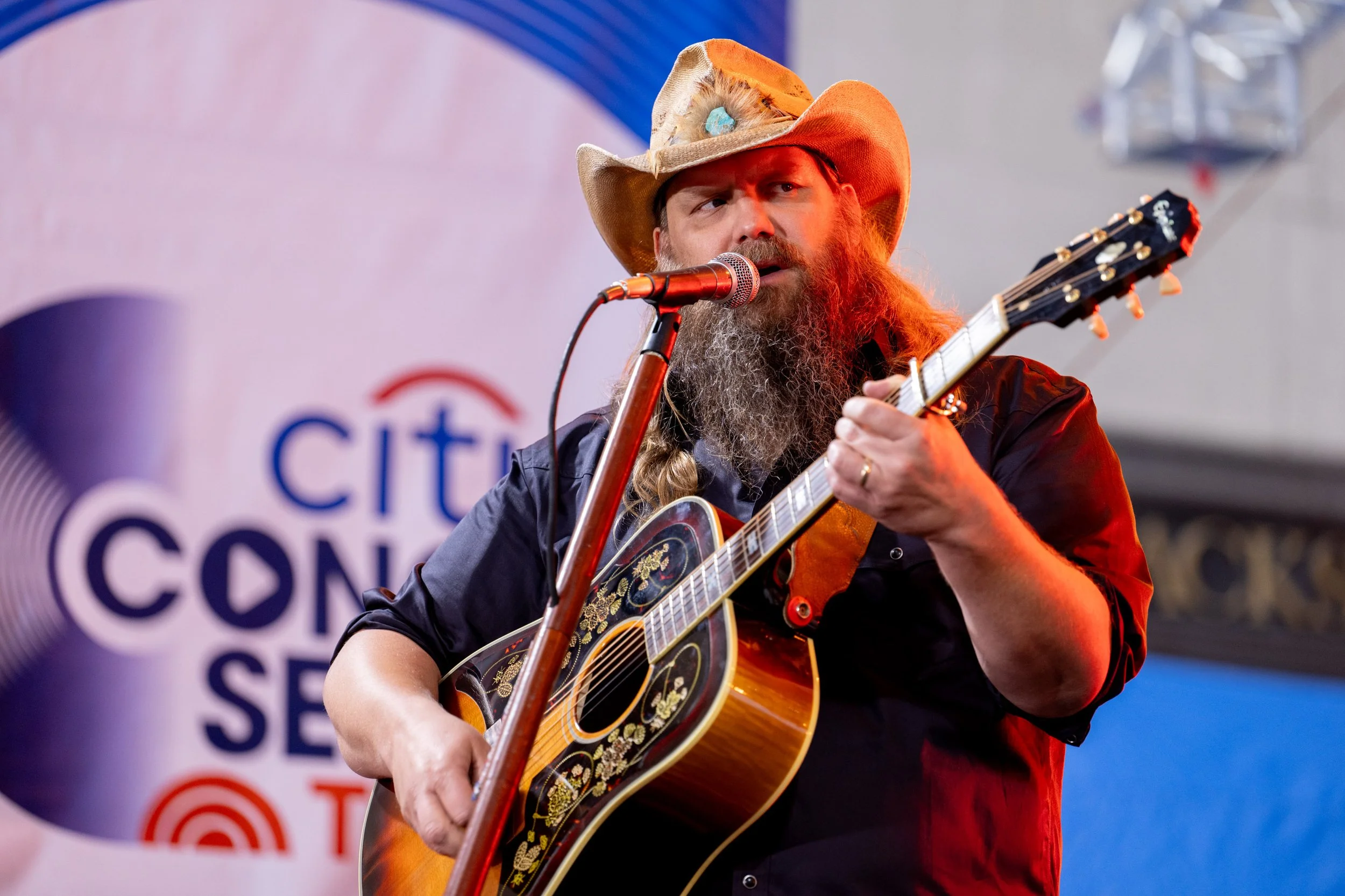 A man with a bushy beard wearing a cowboy hat and dark shirt, playing an acoustic guitar and singing into a microphone at a concert.